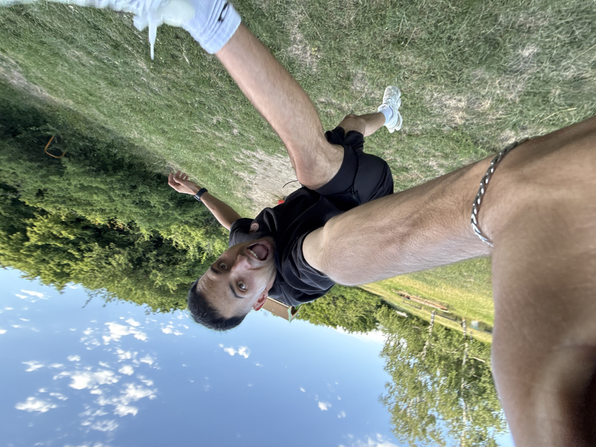 The image shows a first-person perspective of a man in Shumen, Bulgaria, on a grassy outdoor slope under a blue sky with scattered clouds. The man is wearing a black t-shirt, black shorts, and white sneakers. He appears to be in motion, possibly jumping or running, with his mouth open in an excited or surprised expression. One arm is extended forward, holding what appears to be a phone or camera, capturing the selfie. Another arm is raised behind him, and his legs are bent. The background consists of lush green trees and a building with a brown roof in the distance. The overall impression is of a dynamic and energetic moment outdoors.