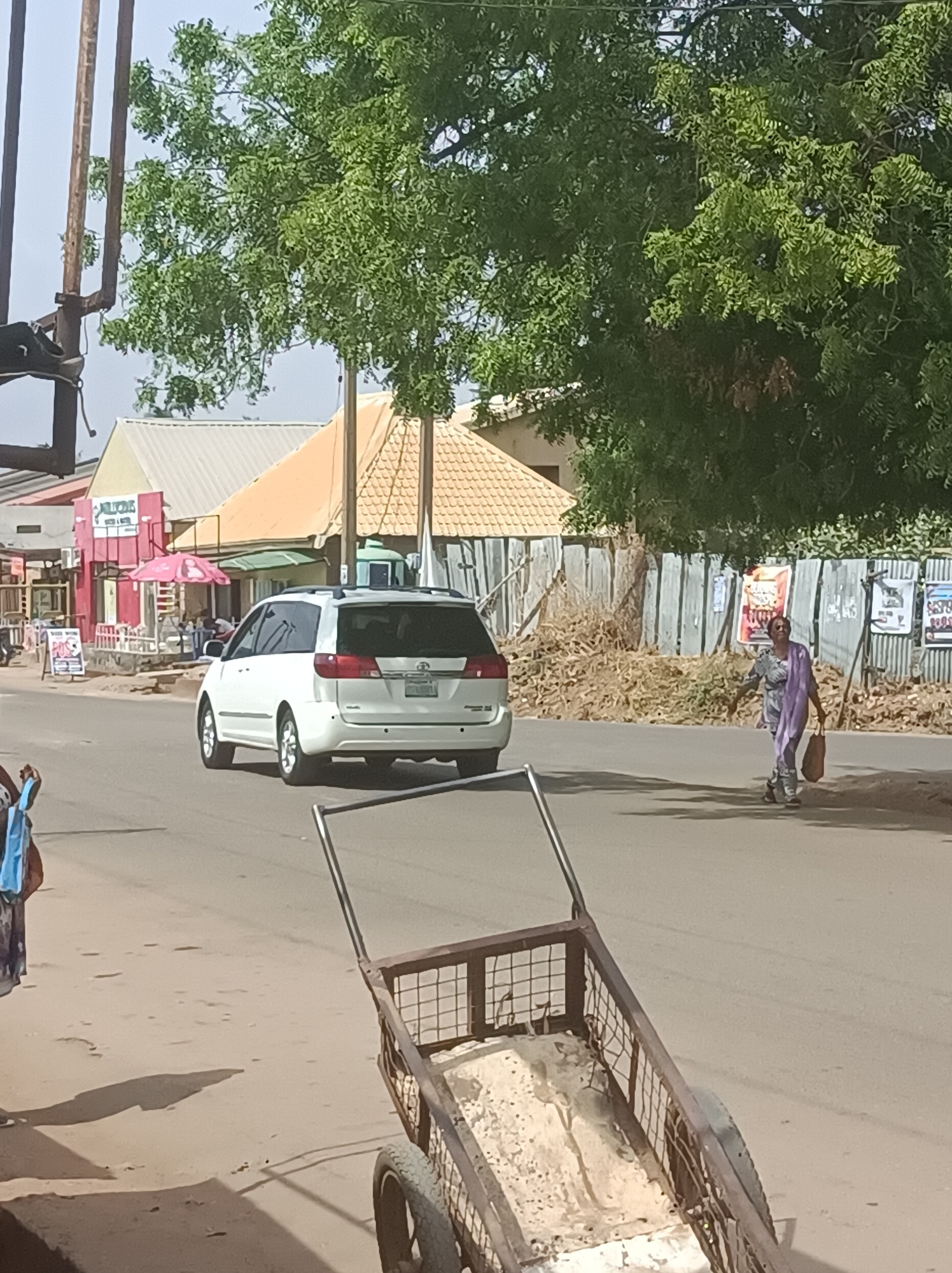 The image depicts a sunlit street scene in Narayi, Nigeria, bustling with a mix of pedestrian and vehicular elements. In the mid-ground, a white Toyota Sienna minivan is either driving or parked on the asphalt road, seen from its rear. The vehicle has roof rails, and its license plate contains partially discernible text that could read "LAGOS".
To the right of the minivan, a woman is walking across the road, heading towards the right edge of the frame. She is dressed in a patterned light-colored outfit, complemented by a purple head covering or shawl, and carries a light brown bag in her left hand. Her shadow stretches out behind her on the road.
On the left side of the road, behind the minivan, several buildings are visible. One prominent establishment features a red facade with the text "JILLICHS" and what appears to be "FAST FOOD" or "RESTAURANT" above its entrance. A pink outdoor umbrella and seating areas are set up in front. A white signboard nearby displays "POS" along with other unreadable text. A person with a blue bag is partially visible on the far left.
The immediate foreground is occupied by a rusty metal pushcart or trolley with two wheels, situated on the sandy shoulder of the road. Its interior appears worn and dirty.
On the right side of the road, a corrugated metal fence stretches along, adorned with multiple posters and advertisements, the text of which is largely illegible. Piles of dry brush or refuse are visible behind the fence, with large green trees dominating the upper right portion of the frame. The overall atmosphere is bright and sunny under a clear sky, with strong shadows indicating daylight hours.