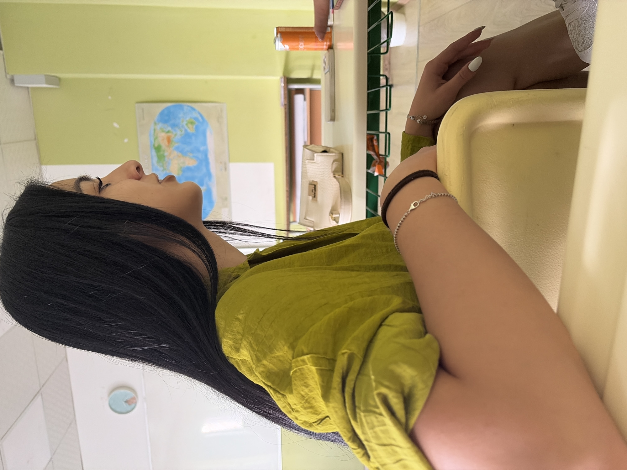 The image is presented on its side, featuring a young woman with long, dark, straight hair, looking upwards and towards the top of the frame. She wears a lime green, loose-fitting top and has a neutral expression.
Her left arm, adorned with a black cord bracelet and a thin silver bracelet, is visible in the lower center. Her right hand, featuring light-colored nail polish and another silver bracelet, is positioned near the top right. A smooth, light yellow surface, possibly a table or tray, occupies the bottom right of the frame. Behind this, parts of another person's arm and leg, dressed in white lace fabric, are visible.
The background primarily consists of a light green wall extending across the upper portion of the frame. A world map is affixed to this wall. Further right, a white wall section contains a small, round analog clock with a light blue face and an orange hand. On the far left, a light green shelving unit holds a white handbag, an orange soda can, and a white remote control. A doorway or corridor is visible beyond the shelving.
The setting appears to be an indoor common area or classroom, typical of a public or educational building, possibly located in Burgas, Bulgaria. The woman seems to be in a moment of rest or contemplation. The scene is brightly lit, indicating daytime indoors. No discernible text is clearly visible.