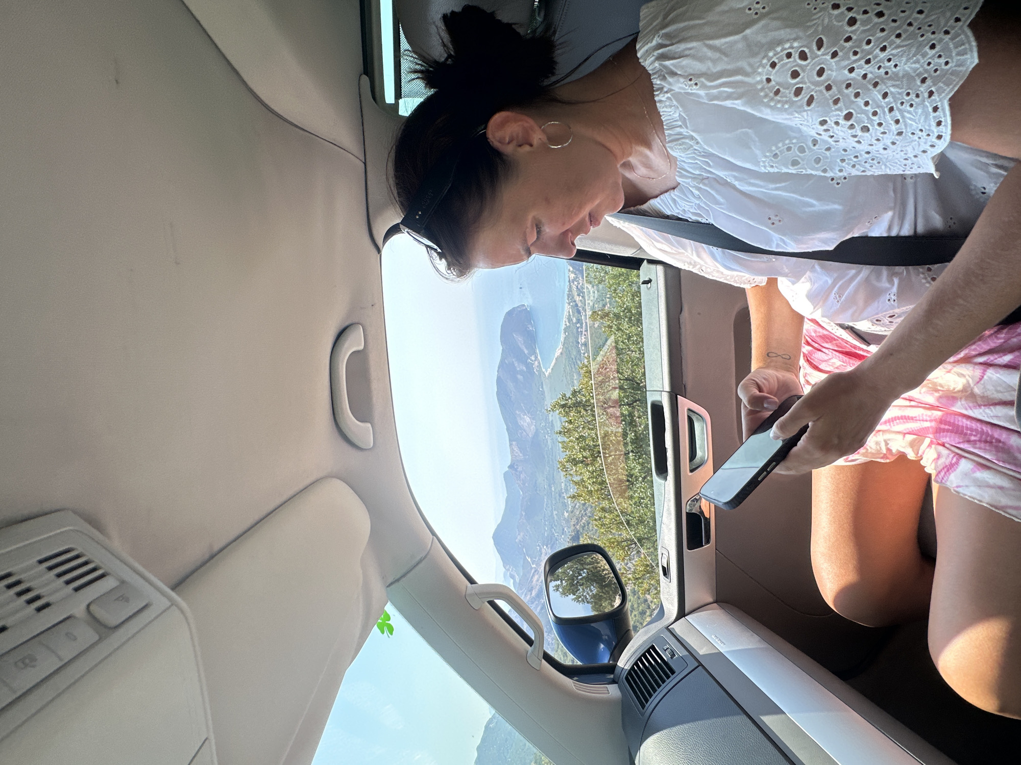 A woman is sitting in the passenger seat of a car, looking at her phone. The car is on a road with a view of the sea and mountains. The woman is wearing a white off-the-shoulder top and pink shorts. She has a tattoo on her left arm. The car's interior is visible, including the dashboard and ceiling. The scene appears to be in Novoselje, Montenegro, based on the landscape.