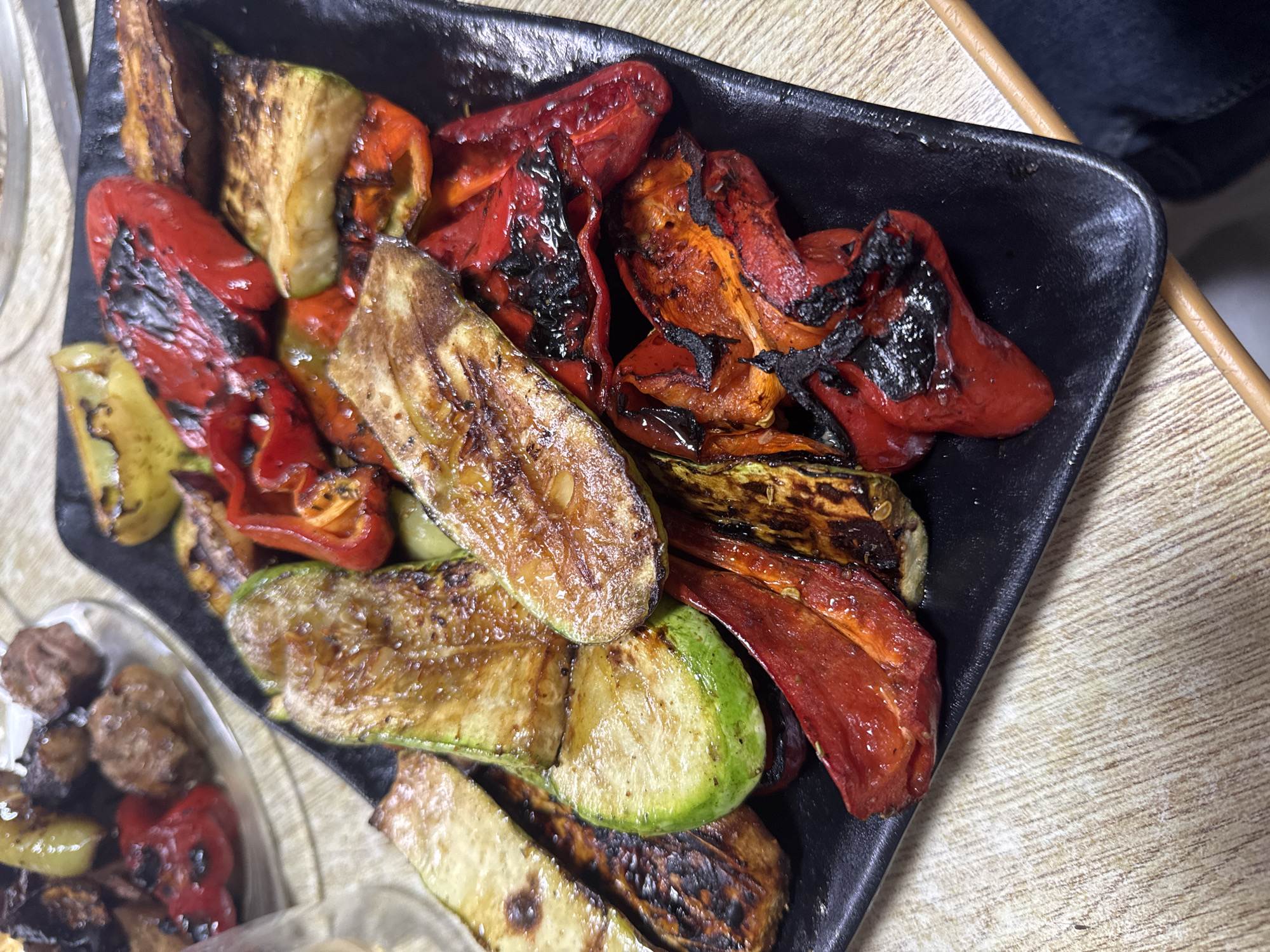 An overhead shot shows a black rectangular plate filled with grilled vegetables, including zucchini, bell peppers, and eggplant, artfully arranged on a wooden table. The vegetables are slightly charred in places, indicating they have been grilled to perfection. In the background, a plate of grilled meat is partially visible, suggesting a hearty meal. The overall impression is one of a delicious and satisfying meal, prepared with care and presented in a rustic setting.