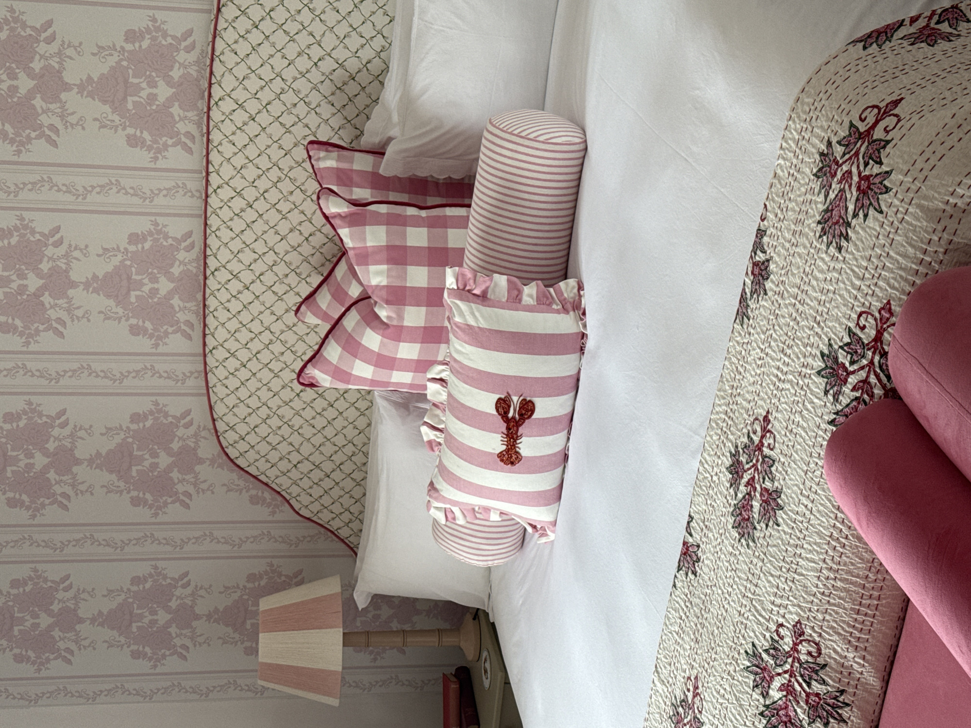 The image presents an overhead view of a bedroom. On the left, wallpaper with a repeating floral motif in light pink covers the wall. A bedside table with a pink-striped lampshade and two small books sits next to the bed.
The bed itself features a variety of decorative pillows in pink and white patterns, including stripes, gingham, and a pillow adorned with a red lobster. A white duvet is neatly spread across the bed. A floral-patterned quilt covers the edge of the bed on the right side. A pink upholstered chaise lounge is partially visible in the bottom right corner.
There is no indication of the location being Cape Town, South Africa, or any people present in the image. The scene depicts a well-decorated and tidy bedroom.