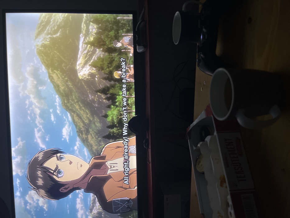 The image depicts an indoor scene in a dimly lit room in Leverkusen, Germany. A large, wall-mounted or stand-supported flat-screen television dominates the upper portion, displaying an anime scene from "Attack on Titan." The central character, Eren Yeager, wearing a brown military-style jacket with the Survey Corps emblem, is visible on the left side of the screen, looking to the right with a determined expression. Subtitles at the bottom of the screen read: "All right, already! Why don't we take a break?" The anime background features a bright blue sky with scattered clouds, verdant mountains, and green trees. Two smaller, similarly uniformed characters are visible in the mid-ground of the anime scene, one appearing to hold a stick or spear.
Beneath the television is a dark wooden shelf or media console, on which a dark, indeterminate object (possibly a remote control) rests. Cables are visible behind this console.
The foreground features a wooden table, partially illuminated, displaying several items. On the left, an open white and red cardboard package labeled "TEARSUTEMENT" contains several individually wrapped items, possibly tea bags or snacks; some appear to have been removed. Two dark grey or black ceramic mugs are present on the table. The mug closer to the viewer contains a dark liquid. A black PlayStation game controller is positioned to the right of the package, behind the closer mug, with a second dark mug behind it. Small, metallic, circular objects are scattered on the table between the package and the controller. The overall impression is of a casual, relaxed setting, likely during evening hours, with an individual engaging in media consumption and refreshments.