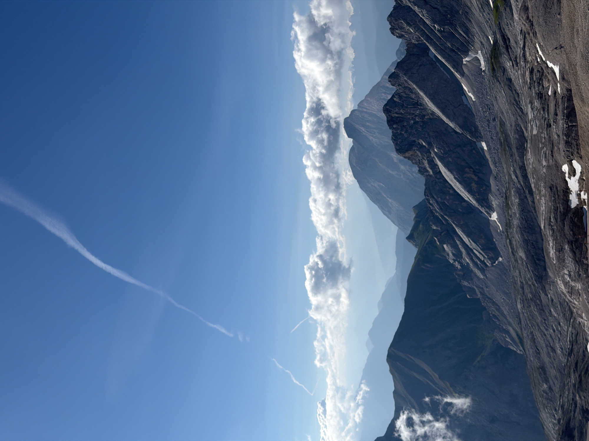 This is a vertical, eye-level, medium shot of a mountain range under a clear blue sky. The sky occupies the top half of the image, with a long, thin white contrail arcing across it from the top left towards the center. In the distance, several layers of mountains fade into a hazy blue. The foreground is dominated by rocky, grey mountain slopes with sparse patches of snow. Some of the slopes are steep and jagged, while others are more gently inclined. White, puffy clouds are visible between the mountain ranges in the mid-ground. There are no people or man-made objects visible in the image. The scene appears to be a natural landscape, likely taken from a high vantage point in the Alps. The location is implied to be near Garmisch-Partenkirchen, Germany, which is known for its mountainous terrain.