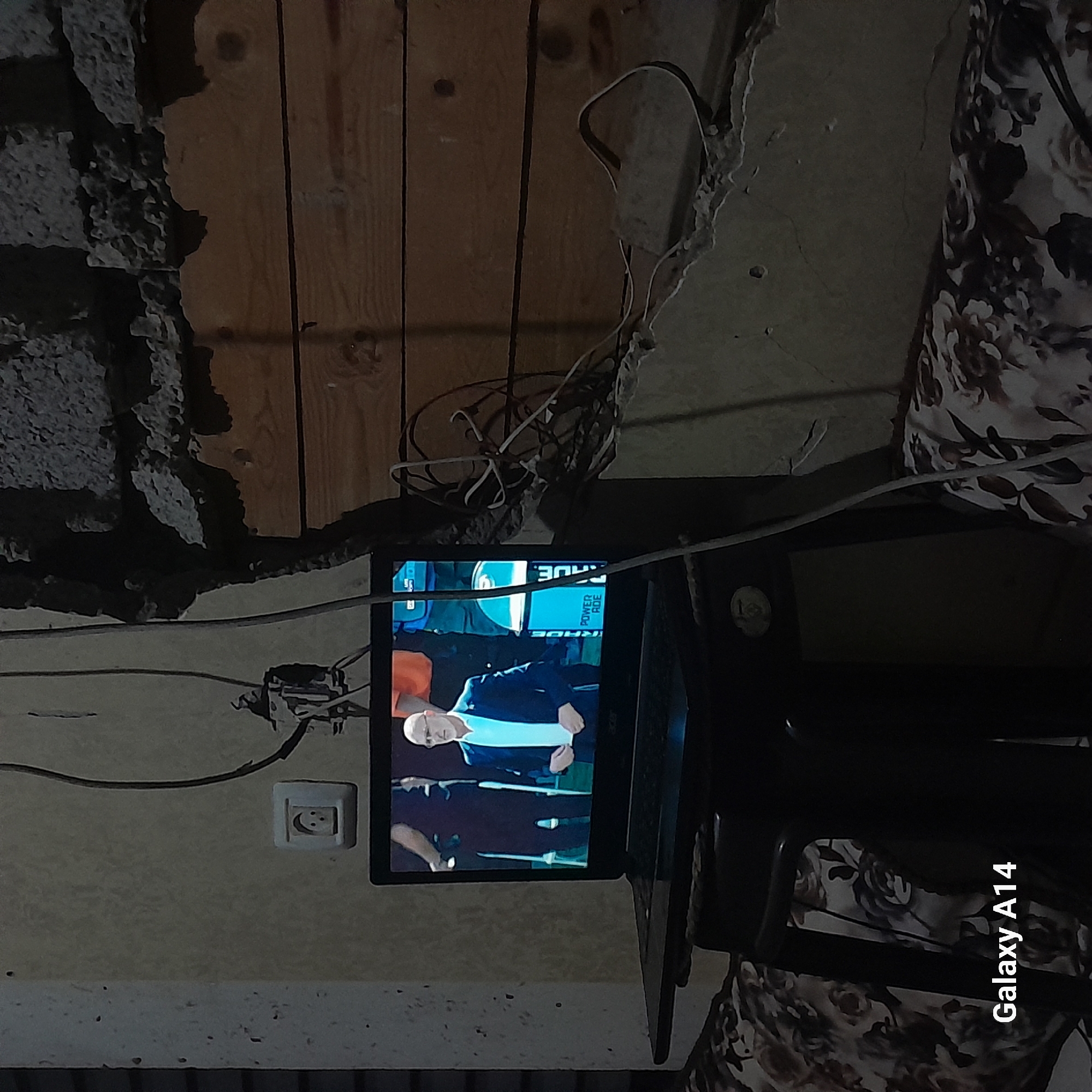 A laptop is placed on a stool, displaying a person in a suit on its screen. The laptop is in a room with damaged walls, showing exposed bricks and wooden planks. Wires are visible, connected to an electrical outlet on the wall. The image also shows a portion of a sofa with a floral pattern. The scene appears to be in Khan Yunis, Palestinian Territories.