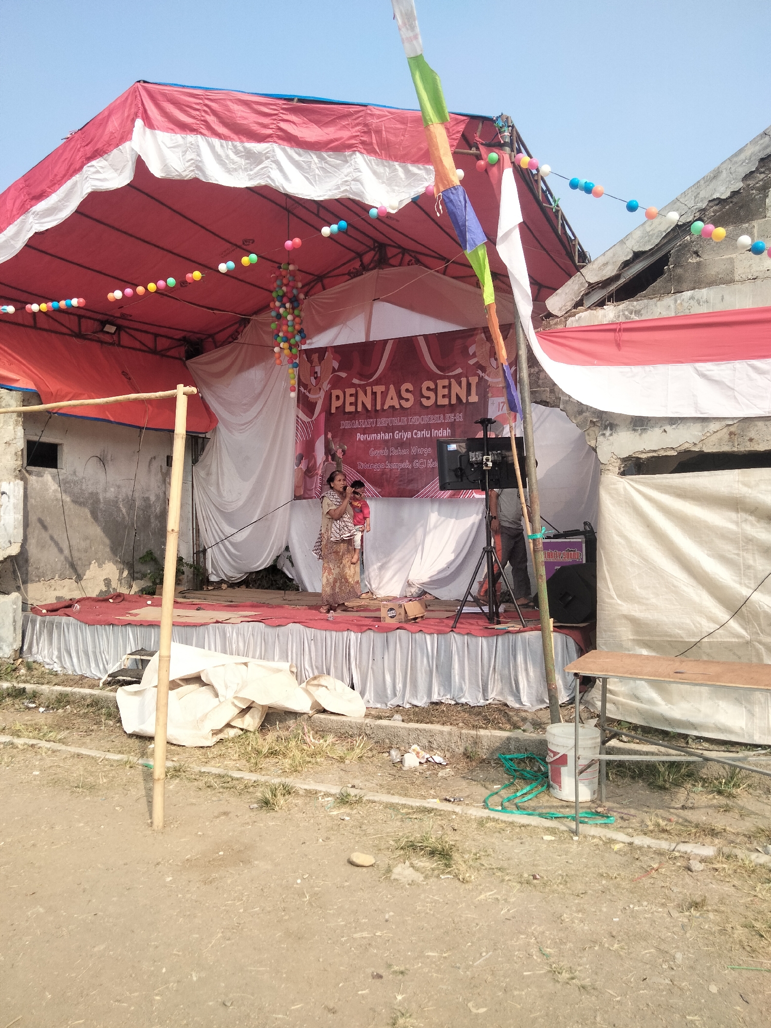 This image captures an outdoor performance stage set up in the city of Cariu, Indonesia. A red and white tent covers the stage, adorned with strings of colorful spherical decorations. A banner displaying "PENTAS SENI" (Art Performance) along with other text is visible behind the performers. On the stage, a woman is holding a child while seemingly singing into a microphone. Another person stands behind a camera setup on the right side of the stage. The environment appears to be a semi-urban or rural area, with a dirt ground and some loosely constructed buildings in the background. There are also various items scattered around the stage, including white fabric drapes, a bucket, and some green hoses.