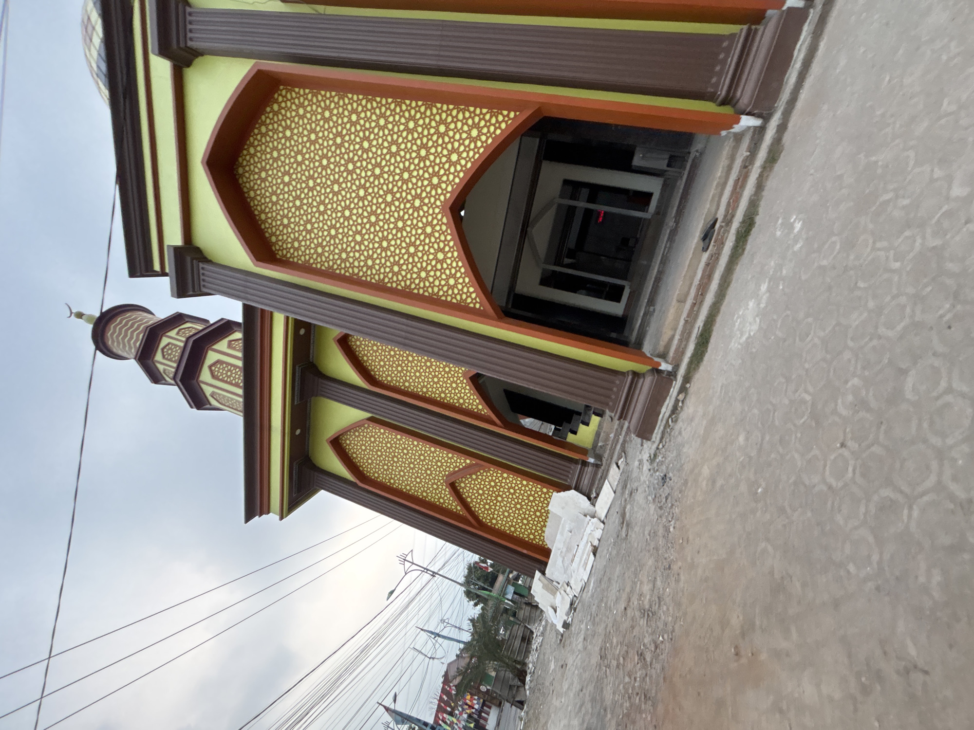The image shows a newly constructed mosque in the city of Bekasi, Indonesia. The mosque has a light green exterior with brown and orange accents. The entrance features large, arched doorways with ornate yellow patterns. A minaret with a pointed dome and decorative bands rises to the left of the main building. The surrounding area appears to be under construction, with piles of white building materials, loose dirt, and gravel on the ground. Electrical wires are visible overhead, indicating proximity to a street. A few flags and a lamppost are also present in the background, suggesting an urban or suburban setting. The sky is overcast.