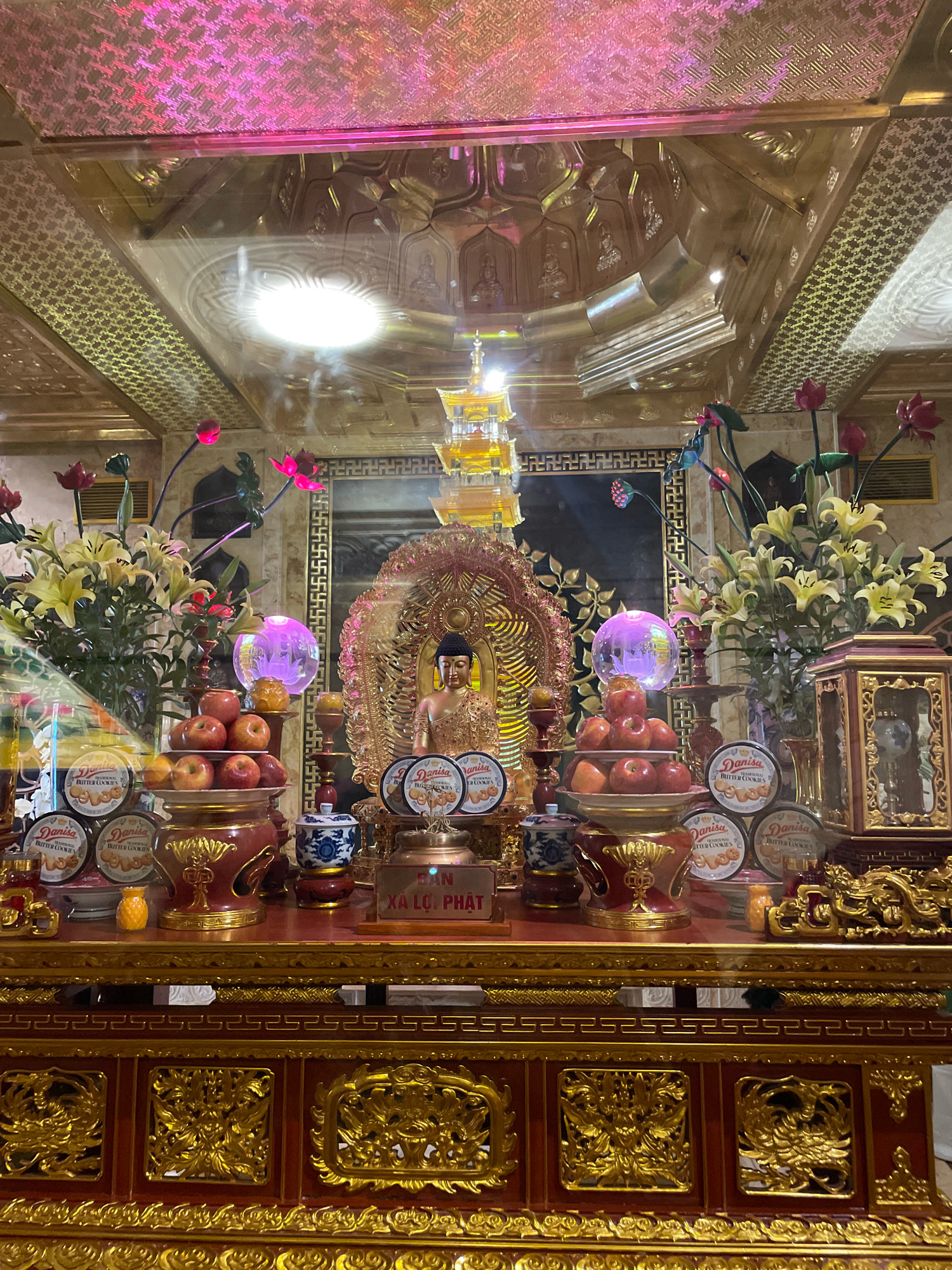 This image captures an altar within a building, likely a temple or a home shrine, in Ta Van, Vietnam. The altar is richly decorated with gold and features a central Buddha statue seated within an ornate, gilded frame. In front of the Buddha, there is a tiered display of red apples. To the left of the Buddha, a bouquet of yellow lilies and red lotus flowers adds a natural element to the scene. The altar is further adorned with several circular tins of "Danisa Traditional Butter Cookies" and two small blue and white porcelain vases. A lit candle and a translucent sphere emitting a soft purple light are also present. The background of the altar includes a golden tiered pagoda structure and patterned wall hangings. The ceiling above the altar is ornate, with gilded carvings and pink lighting, reflecting in the glass enclosure protecting the altar. The lower part of the image shows the detailed woodwork of the altar's base, featuring intricate golden carvings of dragons and floral motifs against a dark red background.