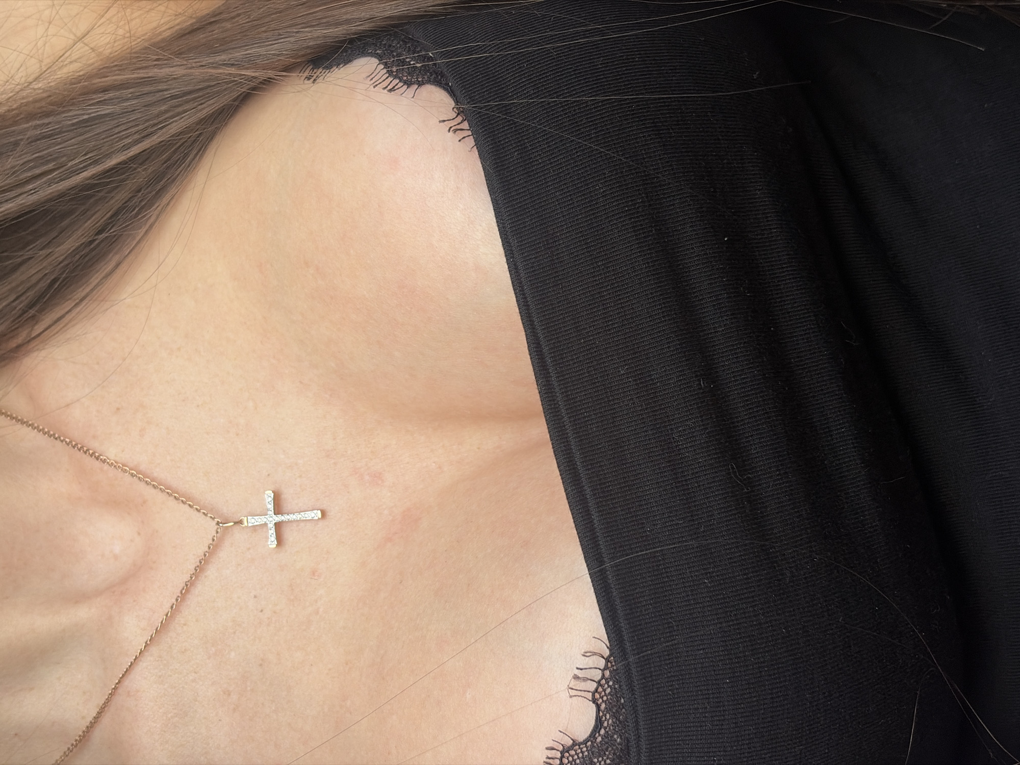 An upper torso image of a person wearing a black garment trimmed with lace. A gold cross pendant hangs from a delicate gold chain around the person's neck. The person has brown hair, partially visible in the upper left. The scene's location within Saint Petersburg, Russia, cannot be determined from the provided image.