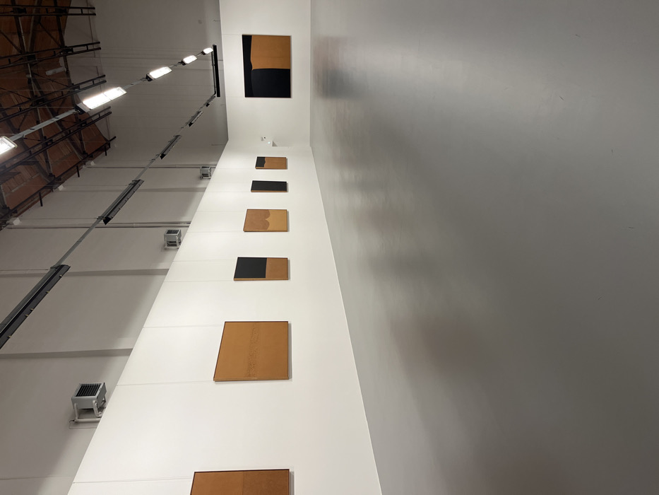 The image captures an interior view of a large, high-ceilinged space, likely an art gallery or exhibition hall. The perspective is looking upwards along a tall, white wall that serves as a display surface for a series of artworks.
On the left side of the frame, several prominent overhead light fixtures are visible, extending in a linear fashion across the high ceiling. These fixtures appear to be long, rectangular units with multiple bright bulbs providing ample illumination. Below these lights, a darker, exposed structural ceiling or series of beams can be seen, hinting at an industrial or minimalist architectural style. Further down the white wall, several square-shaped vents or smaller lighting units are mounted.
The main focal point is the vertical arrangement of seven rectangular artworks displayed along the pristine white wall. These pieces primarily feature an earthy, light brown or tan color, possibly resembling wood, cork, or a natural fabric, often contrasted with sections of solid black.
Starting from the top and moving downwards:
1. The topmost visible artwork is a large, tall rectangle, predominantly tan with a significant black section at the bottom.
2. Below it is a smaller, horizontal rectangular piece, also tan with a black bottom section.
3. The third piece is a small, horizontal rectangle, entirely black.
4. The fourth artwork is a medium-sized, horizontal tan rectangle with a subtly curved bottom edge, giving it a slightly organic shape.
5. Following that is another medium, horizontal tan rectangle with a black section covering its right side.
6. The sixth piece is a larger, squarish tan rectangle, featuring subtle vertical textural lines or markings.
7. The seventh and lowest visible artwork is a horizontal tan rectangle, partially cropped at the bottom of the frame.
All artworks are hung with thin, almost invisible wires, maintaining a clean and minimalist presentation against the white background. To the right of the wall displaying the art, another wall appears in a lighter gray tone, reflecting some ambient light and exhibiting subtle shadows and texture, suggesting a wider space. The overall impression is one of modern aesthetics, focusing on form, color, and texture within a well-lit, expansive environment. No people or visible text are present in the image.