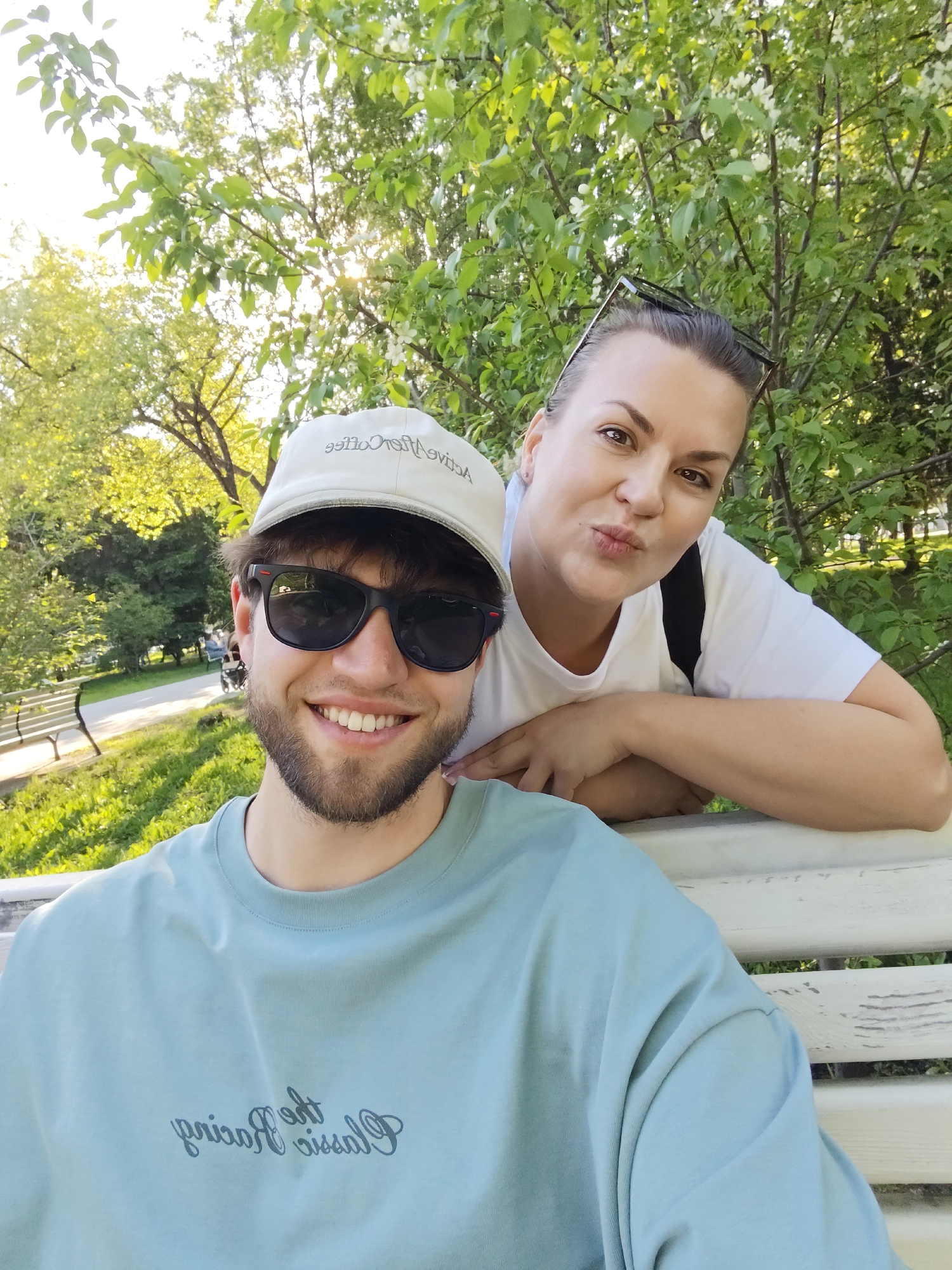 The image shows a selfie of two people sitting on a white park bench. A young man with a beard and sunglasses is in the foreground, smiling. He wears a light blue t-shirt and a baseball cap. A woman is behind him to the right, leaning on the bench with a pouting expression. She wears a white t-shirt and has sunglasses perched on her head. The background features a park with trees and grass. The image was taken in the city of Novosibirsk, Russia.