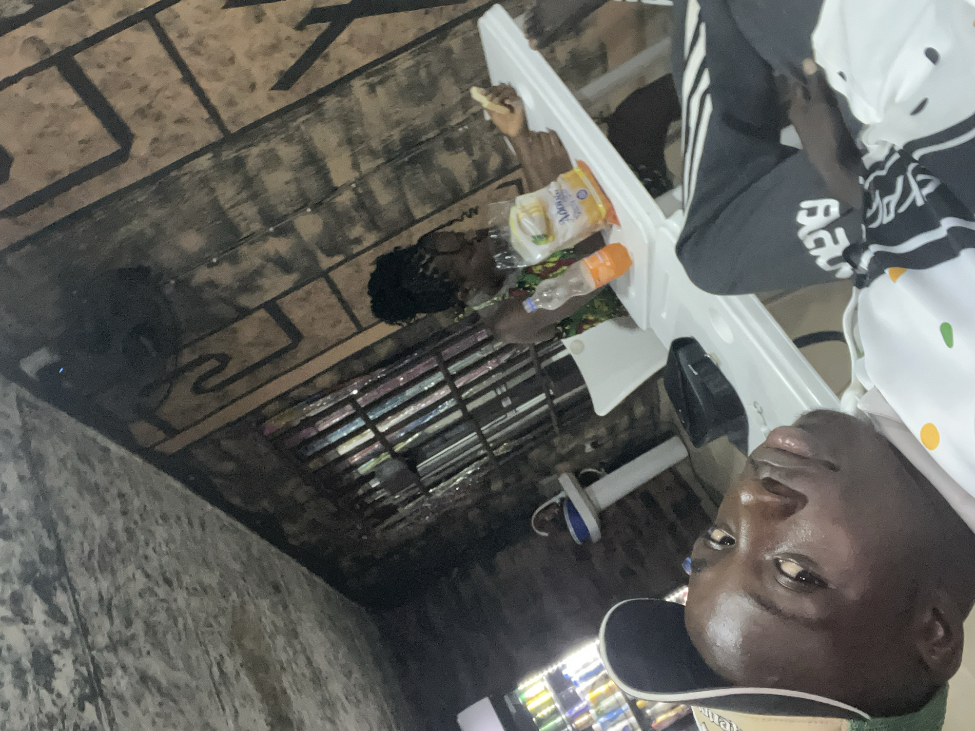 In Nkpor, Nigeria, a close-up, low-angle shot focuses on a young Black man wearing a baseball cap and a white shirt with colorful dots. He is lying down, looking upwards with his head tilted. In the background, a Black woman with her hair in braids sits at a white table, holding a piece of food. On the table are a bottle of orange drink and a packaged snack. The environment appears to be an indoor establishment with a textured, possibly concrete or stone wall behind the table, adorned with geometric patterns. A white sink and a drinks cooler with colorful bottles are also visible in the mid-ground. The lighting is dim, with a cool tone.