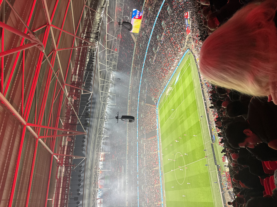The image is taken from a high vantage point inside Wembley Stadium, located in Wembley, United Kingdom, at coordinates 51.558 latitude and -0.28 longitude. The image captures a football match in progress on the pitch below. The stadium is packed with spectators, many wearing red and white. The pitch is green, and a light blue scoreboard is visible in the background. The image shows a group of people in the stands, including a woman with blonde hair, a man with dark hair, and a group of people in the background. The image shows the stadium's roof, which is a complex metal structure with red beams. The stadium lights are on, illuminating the pitch. The image captures the atmosphere of a football match.