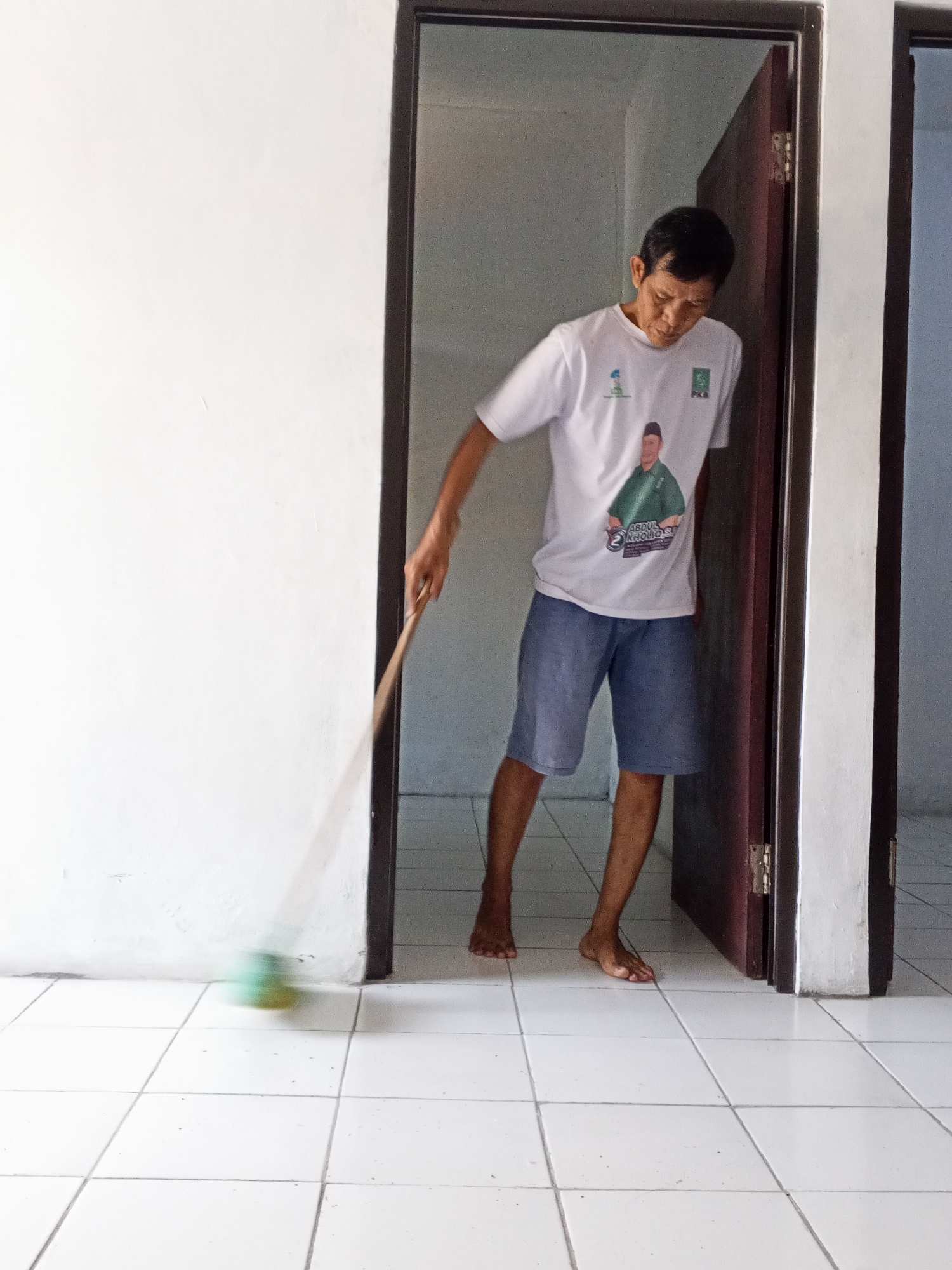 An Indonesian man sweeps a tiled floor with a broom. He is standing in a doorway, wearing a white t-shirt with a picture of a man in a green shirt and blue shorts, and blue shorts. The room is sparsely furnished with white walls and white tiled floors. The man is barefoot. The scene takes place in the city of Ciruas, Indonesia.