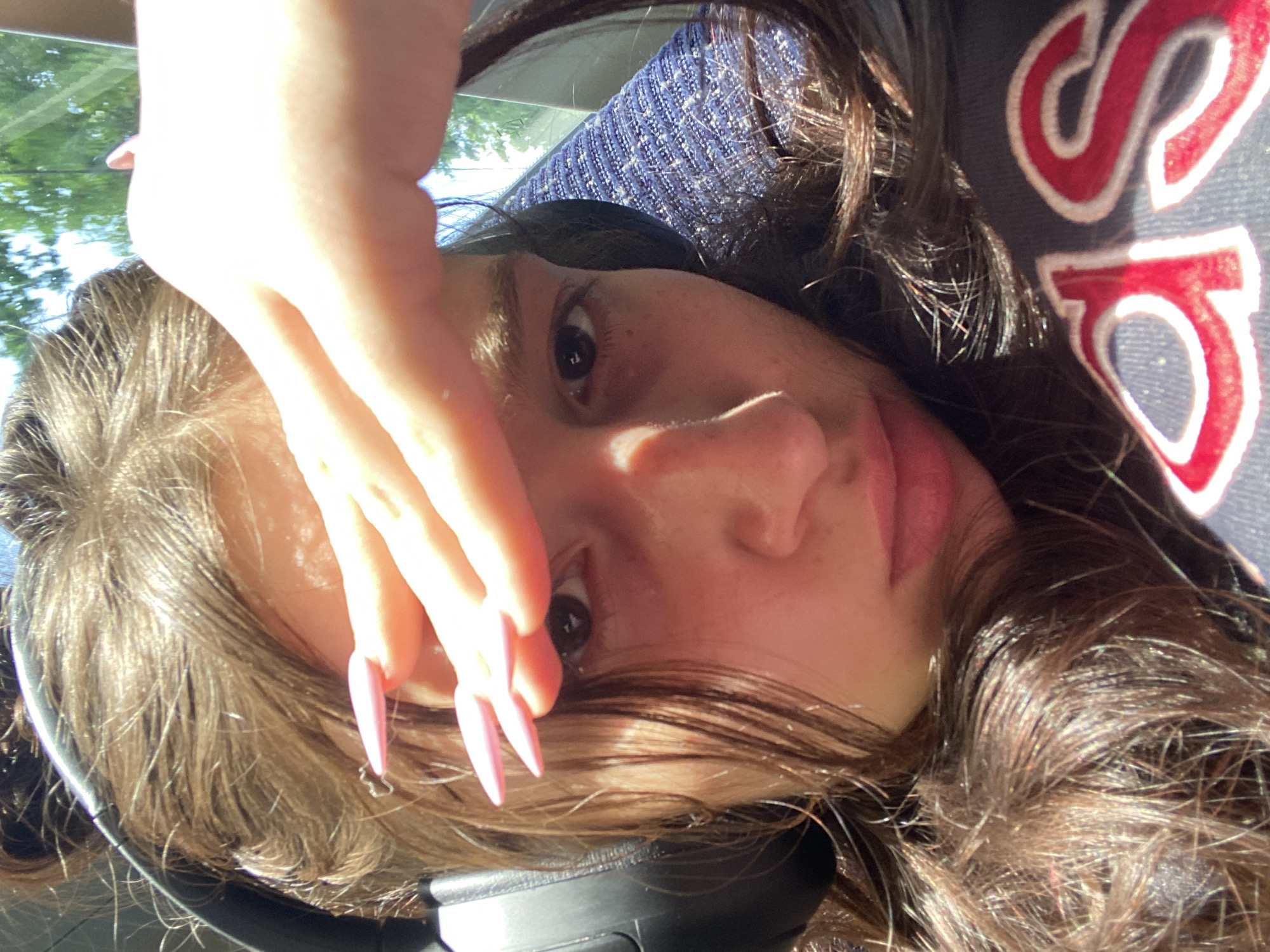 A selfie of a young woman with dark brown hair and fair skin, wearing headphones and a dark blue top with a red and white design. Her left hand is raised, partially obscuring her face, with long, pale pink nails. The background shows a blurred view of trees and a blue textured surface, possibly a seat. Based on the available information and image context, I cannot confidently determine that the location is in the city of London, United Kingdom.