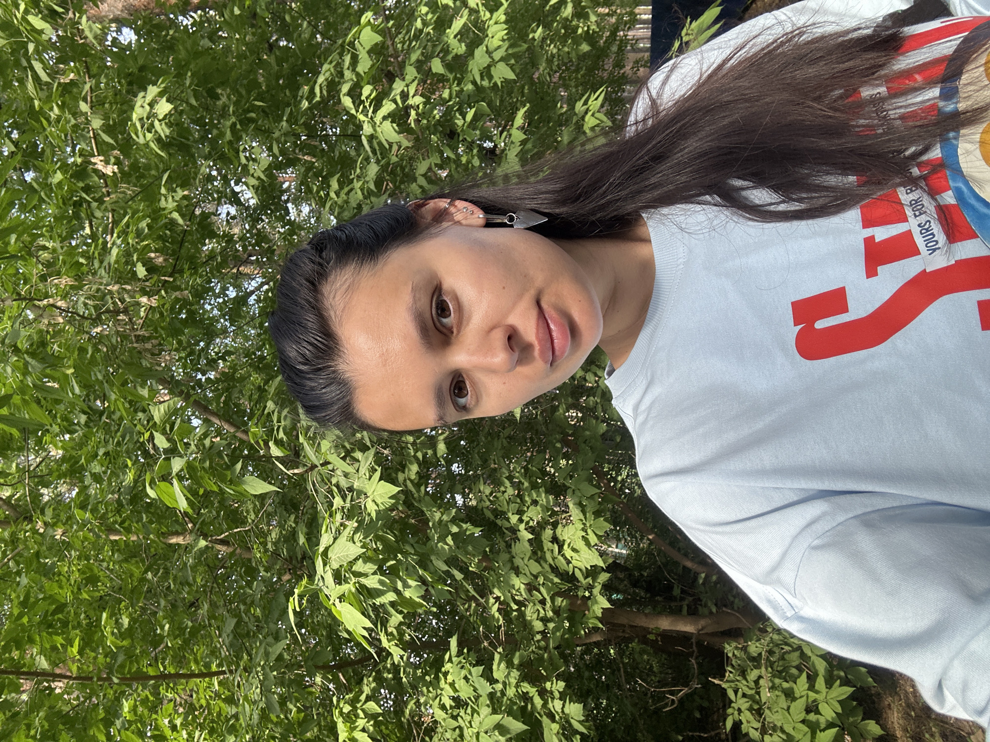 The photograph depicts a woman with long, dark hair standing outdoors. She is wearing a light blue t-shirt with red lettering. She has a silver earring in her ear. The background consists of dense green foliage. The location is indeterminate, and there is no evidence to suggest it is in the city of Chaikovsky, Russia.