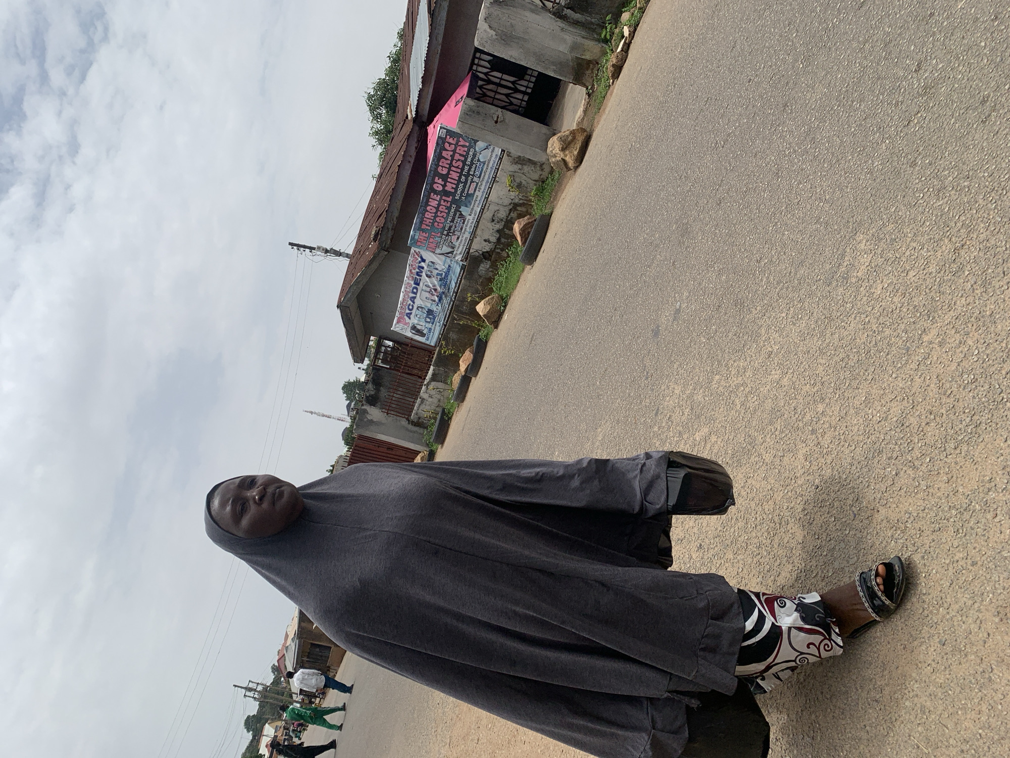 A woman wearing a gray hijab and patterned pants stands on a paved road. In the background, there are buildings, including one with a sign that reads "THE THRONE OF GRACE INTL GOSPEL MINISTRY SCHOOL OF THE SWORD." The sign also mentions "Community Bible Class." Another sign on the building reads "DAYCARS PRECIOUS STONZ ACADEMY." The location appears to be in Jos, Nigeria.