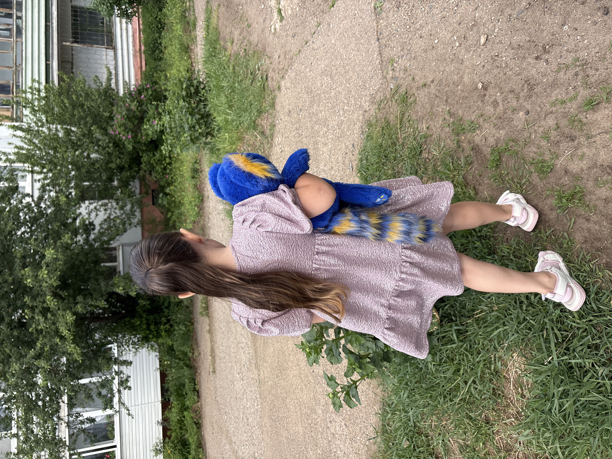 A young girl stands on a dirt and grass path, holding a blue and yellow stuffed animal. She is wearing a light pink dress and white and pink sandals. The background includes a building with white siding and windows, as well as green trees and bushes. The location is potentially Chaikovsky, Russia.