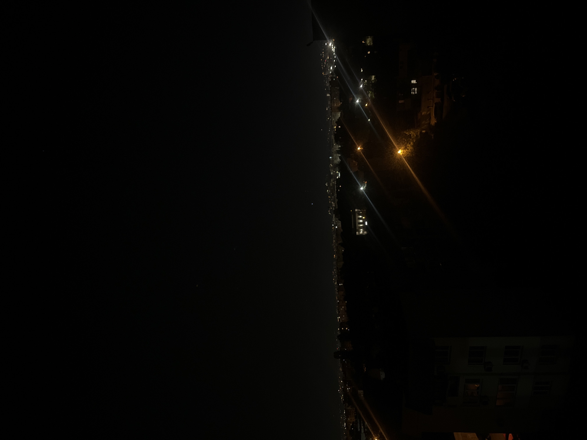 The image shows a night view of Sofia, Bulgaria. The left side of the image is a dark expanse, possibly depicting a river or open space. The right side features a line of streetlights and building lights, indicating a road or urban area. The lights suggest the presence of buildings and vehicles. The environment appears dark and illuminated only by artificial light sources.