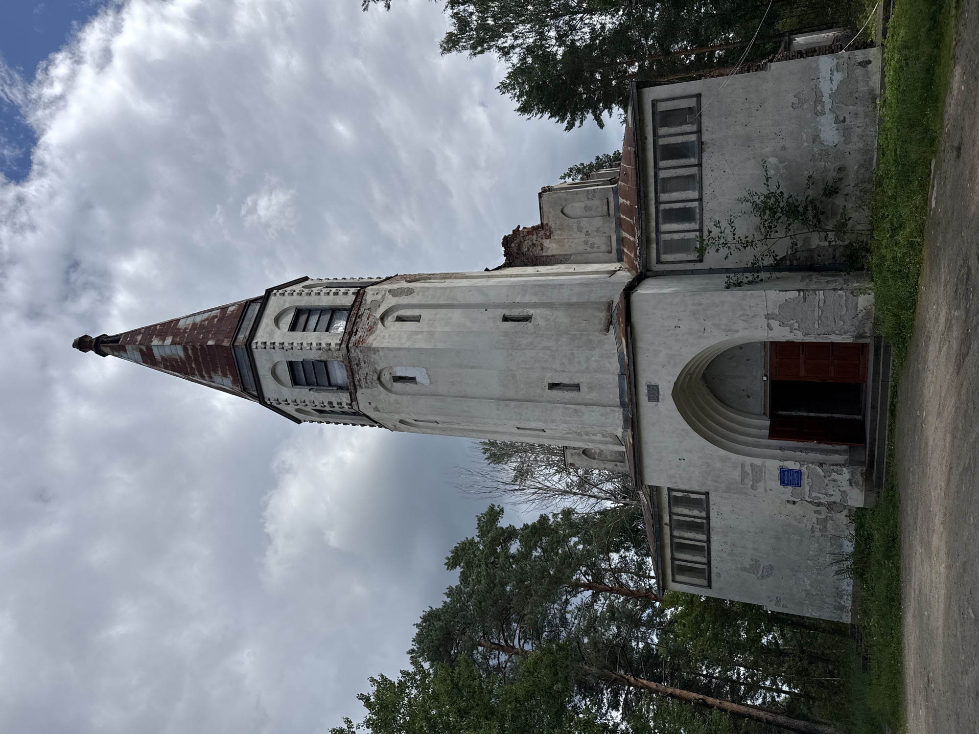 The image shows a light-colored, partially dilapidated church with a tall, narrow tower topped by a rusted metal spire. The church features arched doorways and windows, some of which are boarded up or broken. The exterior walls are peeling and show signs of wear and tear, with exposed brickwork visible in some areas. The church is surrounded by tall green trees, with a cloudy sky overhead. The ground in front of the church appears to be a dirt path. A blue plaque is affixed to the wall to the left of the entrance. The building seems to be located in a rural or forested area. This is the Evangelical Lutheran Church of St. John the Baptist in Lakhdenpokhya, Russia.