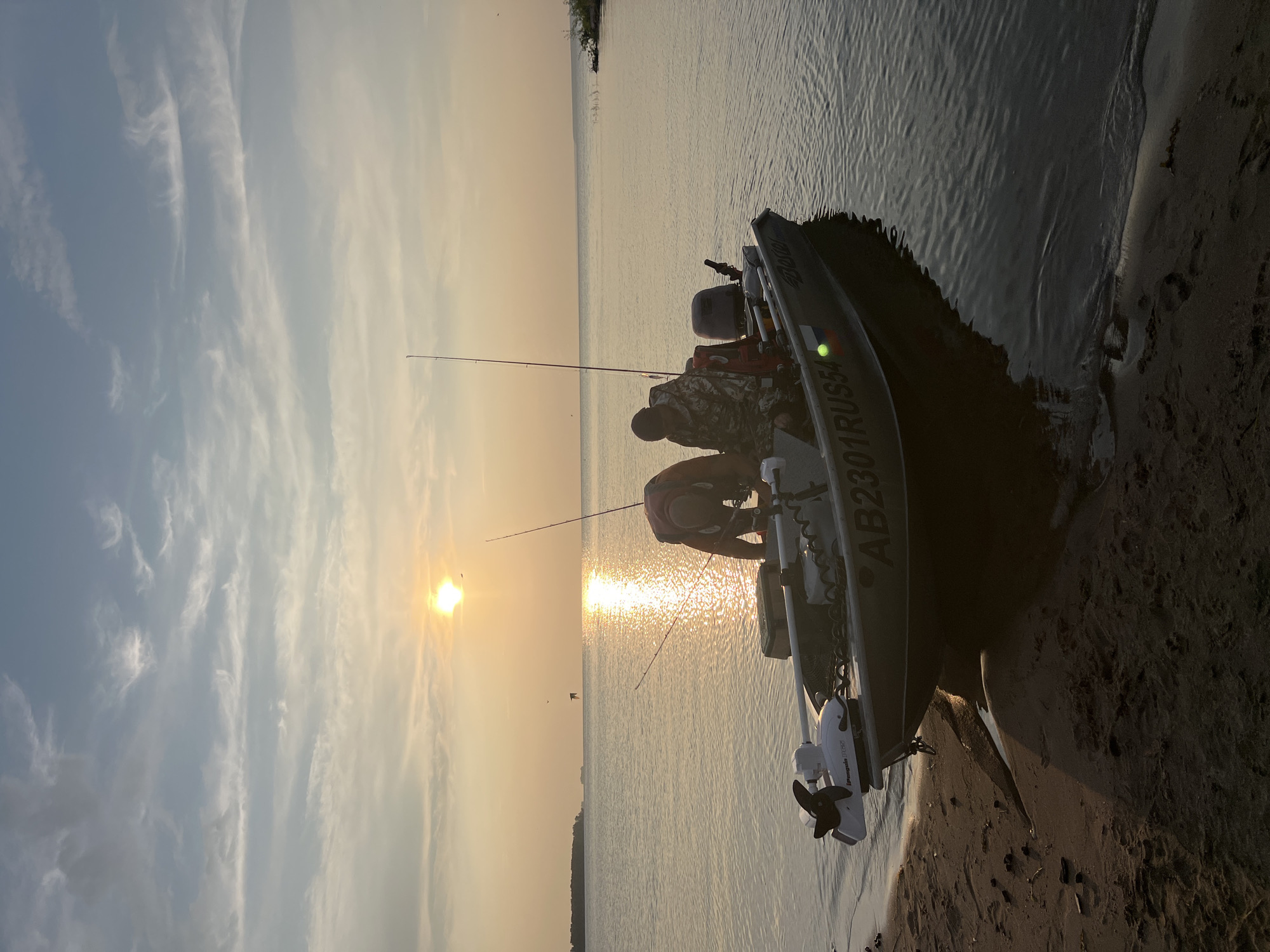 This media file captures a serene moment by the water at sunset. The scene features two individuals engaged in fishing from a boat that is partially grounded on a sandy shore. The boat, with the registration "AB2301RUSSIA" visible, is equipped with a trolling motor. One person is bent over, possibly preparing fishing gear, while the other, wearing camouflage clothing, is also focused on fishing. The sun is low on the horizon, casting a warm, golden glow across the water and sky, suggesting it is either sunrise or sunset. Wispy clouds are scattered across the sky, illuminated by the sun's rays. The water appears calm, with gentle ripples reflecting the light. The overall mood is peaceful and tranquil, indicative of a quiet fishing excursion. The location is suggested to be Borovoe, Russia.