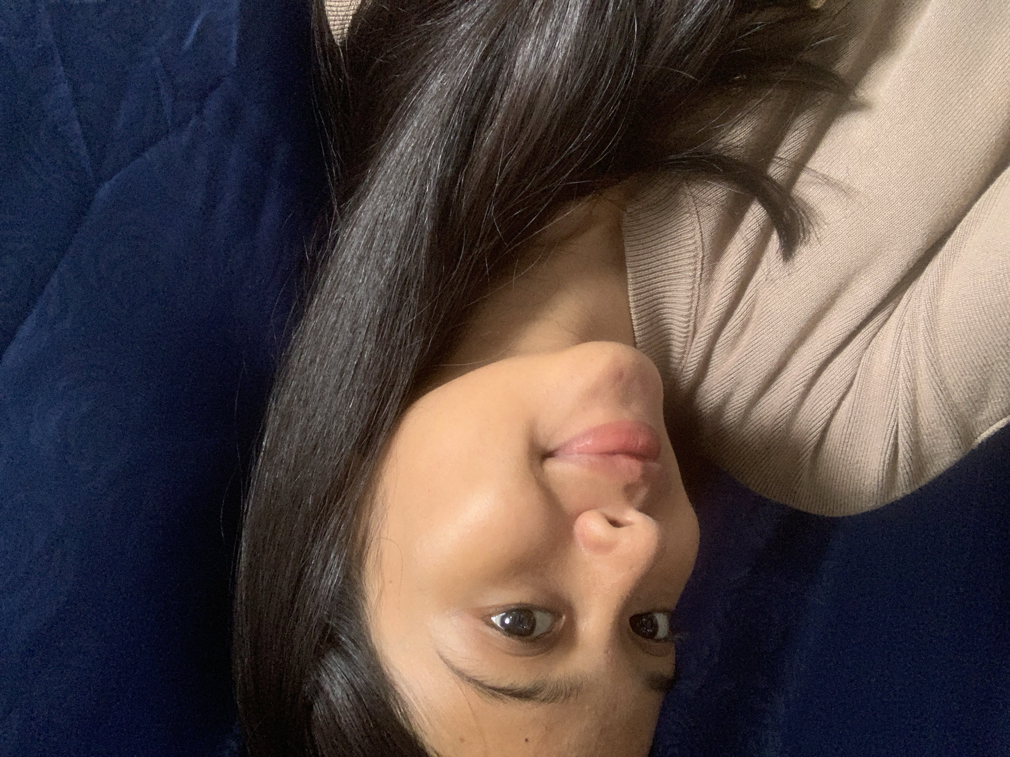 A close-up, overhead shot shows a young woman with dark hair and brown eyes lying on a dark blue surface, possibly a couch or bed. She is wearing a tan, ribbed sweater. Her head is turned to the right, and she is looking towards the camera with a slight smile. Her skin is clear, and her lips are tinted pink. The lighting suggests an indoor environment, with soft illumination highlighting her face and the texture of her clothing. There are no visible contextual clues in the image that would indicate the specific location within Sungai Meriam, Indonesia.