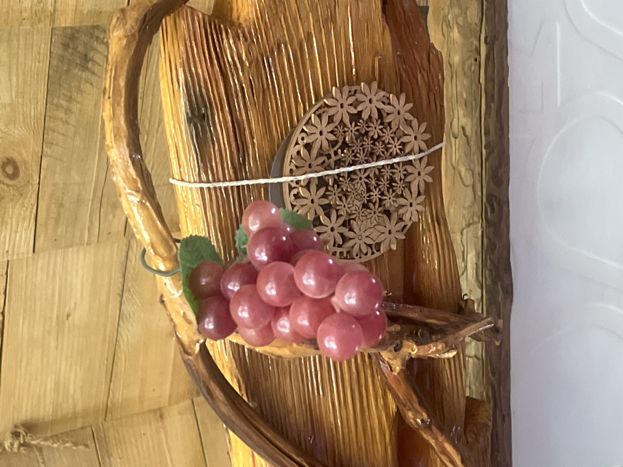 A bunch of artificial red grapes hangs from a twig structure against a wooden background. To the right of the grapes, a circular wooden ornament with a floral pattern is suspended by a white string. The wooden background appears to be made of planks with visible grain and a glossy finish. A rough, textured wooden element forms an arch over the grapes. The bottom of the image shows a white surface with a subtle embossed pattern. The location is Norviliškės, Lithuania.