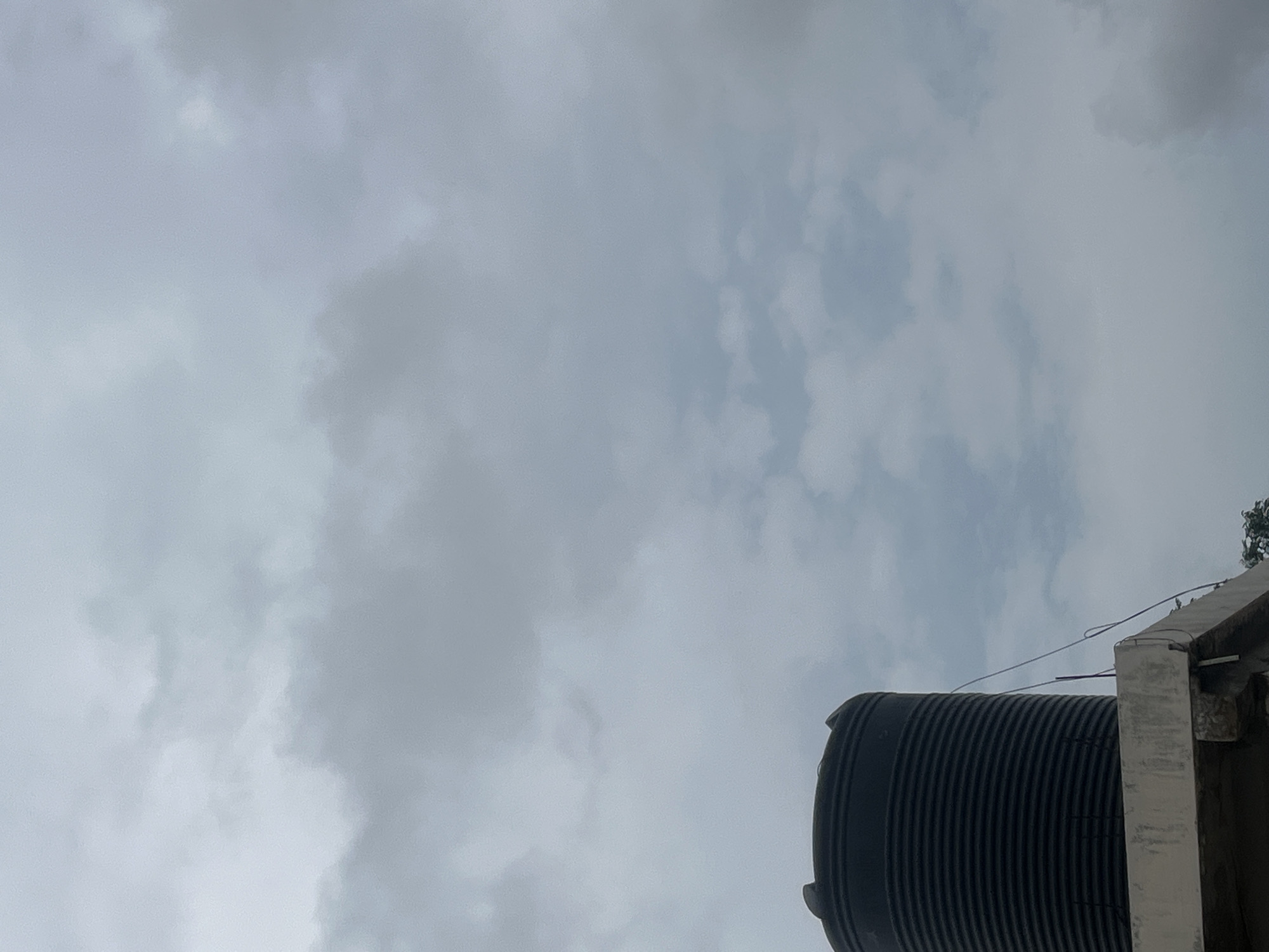 A black, ribbed water tank sits on the edge of a concrete rooftop. The tank has a rounded top with protrusions. A thin wire runs from the tank down the side of the structure. The sky above is overcast with gray and white clouds. The image was taken in Jauro Yinu, Nigeria.