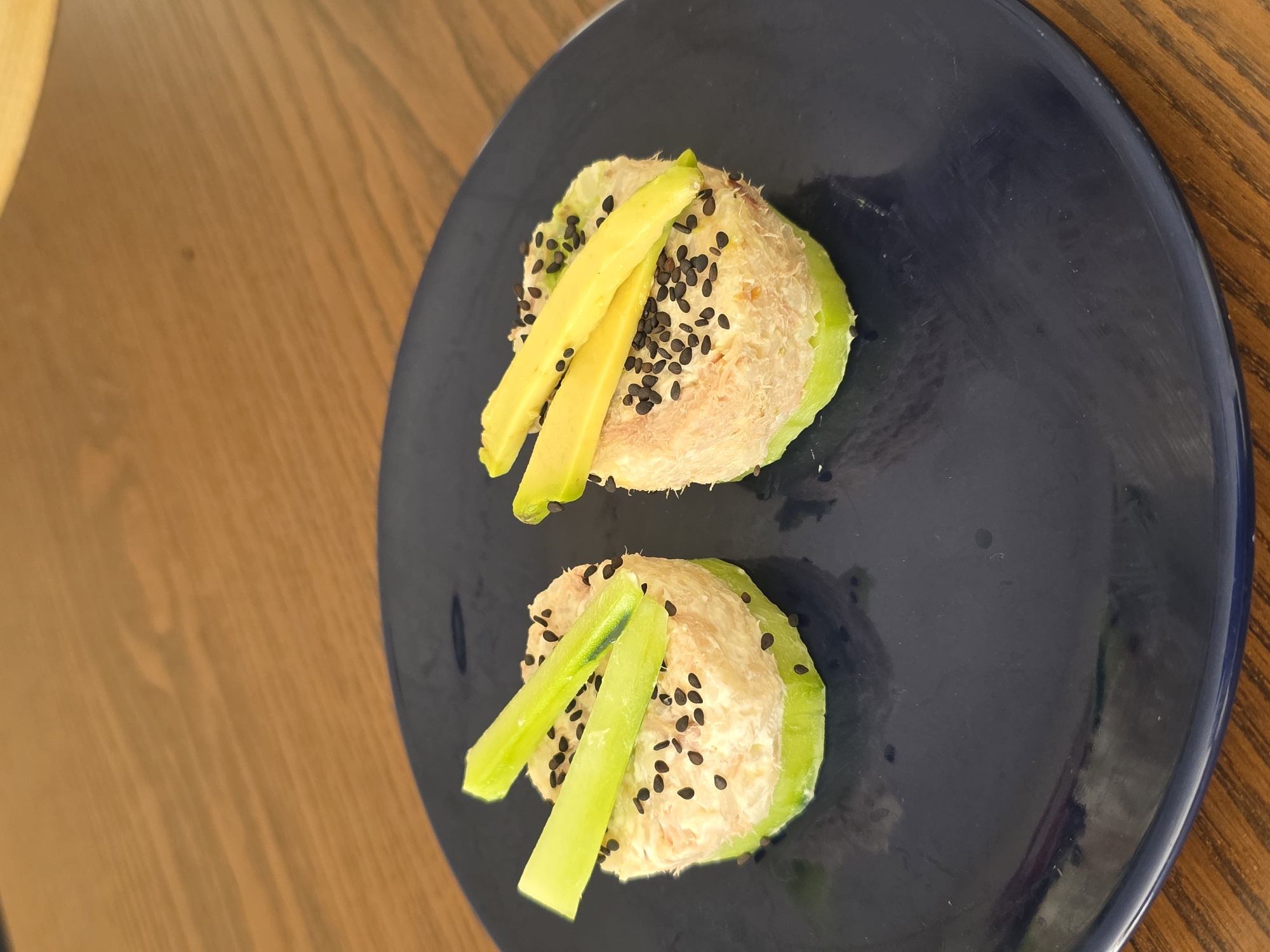 On a blue plate, two mounds of tuna salad, topped with black sesame seeds and avocado or cucumber slices, are placed on a wooden table. The plate is likely indoors, possibly in a restaurant or home. The food appears to be a light lunch or appetizer. The location is not discernible from the image.