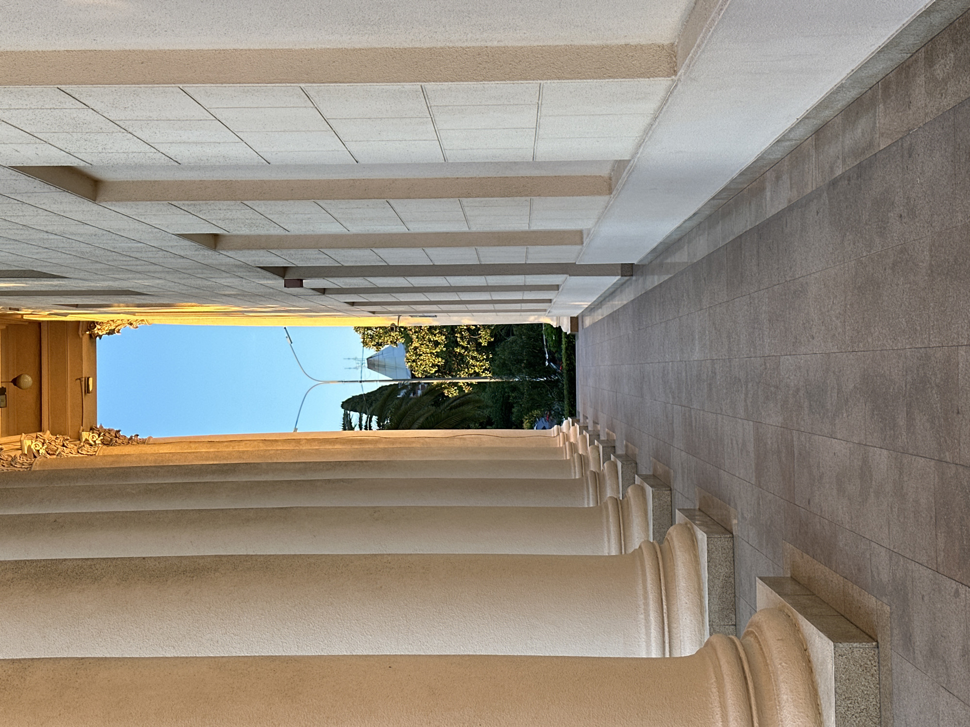 The image displays a long, grand architectural colonnade or portico, viewed from within and looking towards an outdoor scene. When correctly oriented (rotated 90 degrees clockwise), a series of large, light-colored classical columns with rounded shafts recede into the distance on the left side, creating a strong sense of depth. The capital of the foremost column is ornately carved with intricate, possibly gilded, decorative details, and a wall-mounted light fixture is visible above it.
The ceiling above the corridor is composed of textured, light-grey square tiles, intersected by parallel, darker beige beams that create a patterned effect. On the right side, a solid wall constructed from dark grey rectangular tiles runs parallel to the columns, also extending into the distance. Its base features a lighter, speckled grey granite material. The floor of the colonnade is covered with light-colored, rectangular tiles.
The corridor opens up to a bright outdoor environment. A clear, vibrant blue sky is visible at the top of the opening, beneath which lies lush green foliage and trees, including distinct palm trees. In the background, a modern building with a pointed or pyramid-shaped roof can be seen, along with utility lines against the sky.
The scene is brightly illuminated, suggesting daytime under clear weather conditions. The light appears to originate from the open end, casting soft, even light within the corridor, with some warm highlights on the front architectural elements. The absence of people contributes to a serene and expansive atmosphere. The classical architecture combined with subtropical vegetation aligns with the provided context of Sochi, Russia. There is no visible text in the image.