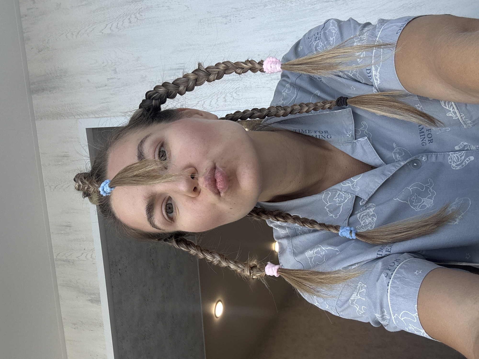 A woman is photographed from below, her face angled toward the camera with pursed lips. She has light brown hair styled in four braids, each secured with colored hair ties: blue, pink, black, and blue again. She wears a light blue pajama top adorned with white cartoon illustrations. The background includes a white, wood-paneled wall in the upper left corner and a gray wall with recessed lighting in the lower left. Location: Unknown, Unknown.