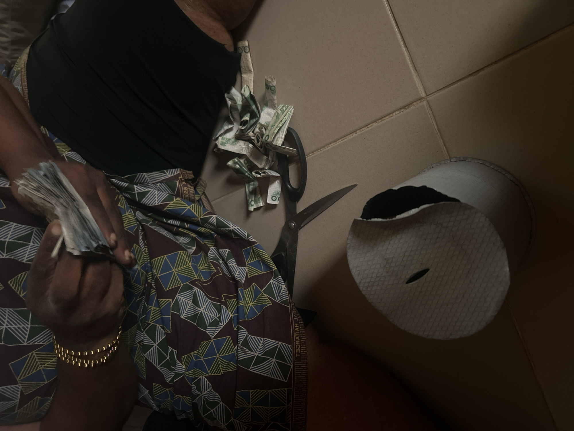 An individual, wearing a black top and a patterned skirt, is holding a stack of banknotes in their left hand. A gold bracelet is visible on their left wrist. Next to the individual are cut-up banknotes and a pair of scissors, suggesting the banknotes were intentionally destroyed. A white hat with a black interior is lying on the tiled floor. The scene is indoors, with tan-colored tiles. The location is Unknown, Unknown.
