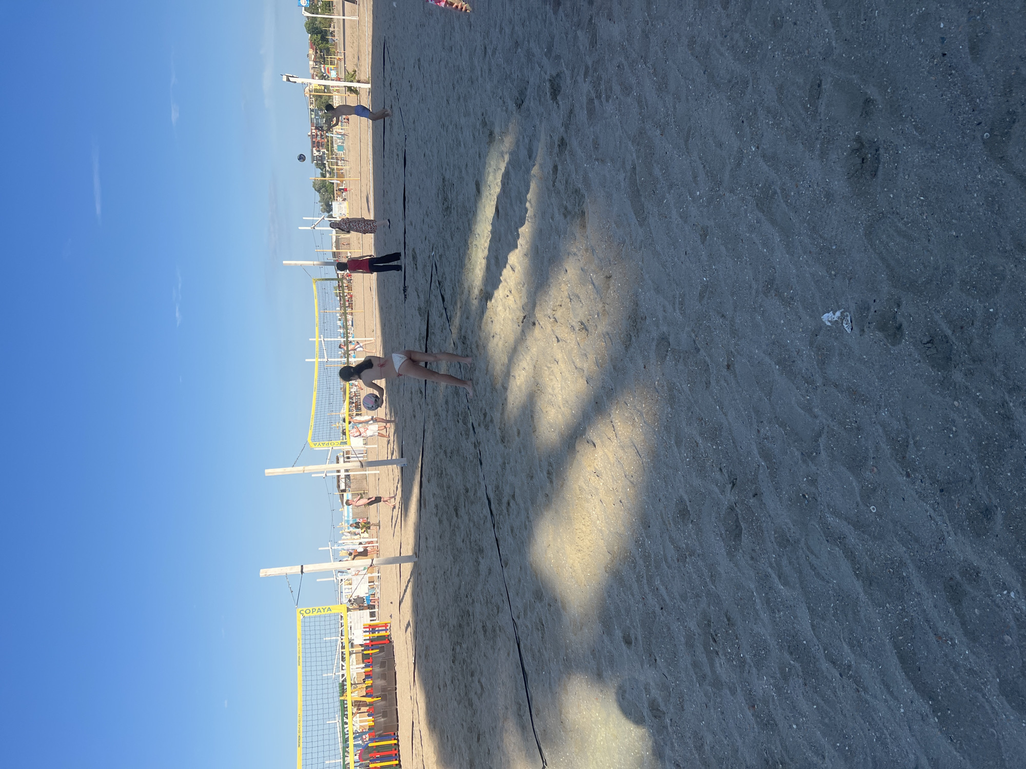 A beach volleyball game is in progress on a sandy beach under a clear blue sky. The scene depicts multiple individuals playing volleyball on a court marked by black lines and a net. In the foreground, a young female player in a white bikini is positioned on the sand, facing away from the camera. In the mid-ground, another young female player is in the process of hitting a volleyball. To her left, two individuals are standing near the net. Further in the background, several other people are scattered across the beach, some watching the game and others engaged in activities. The beach is equipped with volleyball nets and poles, indicating a designated recreational area. The presence of buildings and vegetation in the distance suggests an urban coastal setting. The sandy terrain shows some footprints and shadows cast by the sun. It is a bright, daytime scene.