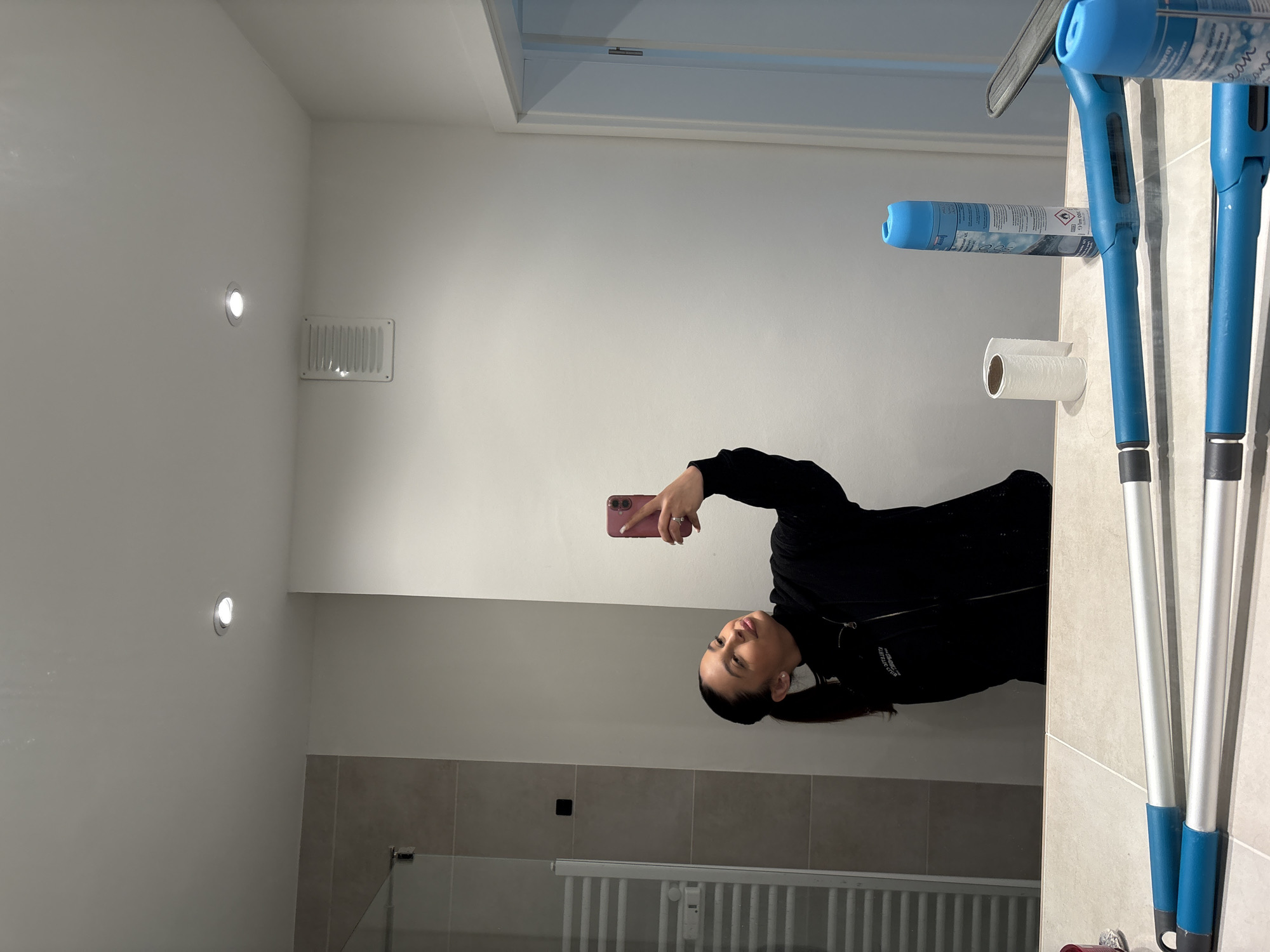 The image depicts a person in a bathroom, taking a selfie in the mirror. The bathroom has white walls and ceiling, with recessed lighting. There's a ventilation grille on the wall. The lower portion of the wall is covered with beige-colored tiles. A white radiator and glass shower enclosure are visible. The mirror reflects the person holding a pink smartphone. Beside the mirror are bathroom cleaning supplies including a blue spray bottle and cleaning tools, alongside a roll of toilet paper. The image does not provide any information to indicate the location as Mönchengladbach, Germany.