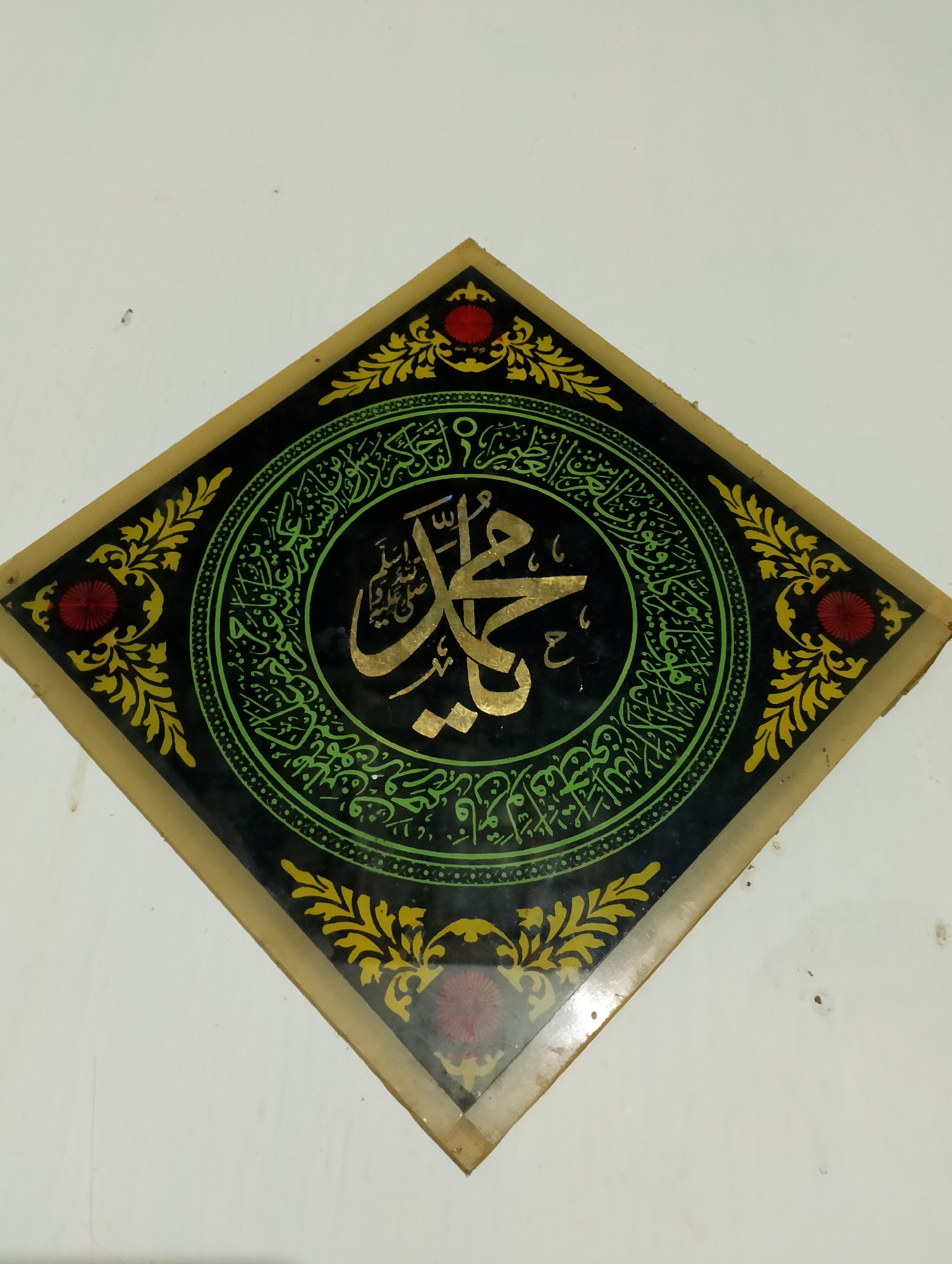 An overhead shot shows a square Islamic calligraphy artwork mounted on a white wall. The artwork is rotated 45 degrees, appearing as a diamond shape. It features a central circular motif containing golden Arabic calligraphy against a black background. This central circle is enclosed by a green, circular pattern of smaller Arabic script and decorative elements. The outer edges of the artwork are black with ornate yellow floral designs and red floral accents at the four corners of the diamond shape. The entire artwork is framed by a golden border. The white wall has a few dark specks, and the lighting is ambient. The artwork is located in the city of Rembang, Indonesia.