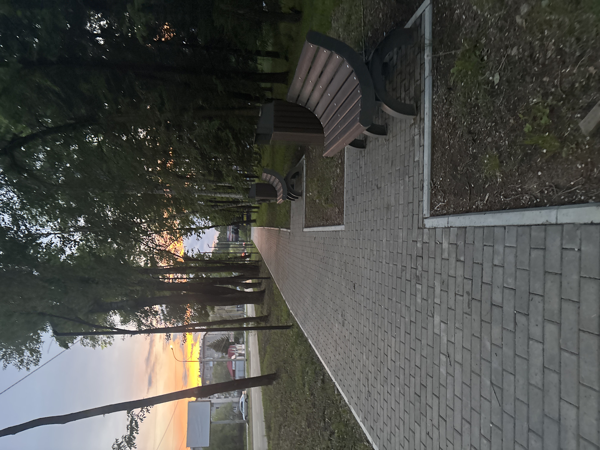 The image presents a tranquil outdoor scene captured during sunset, likely depicting a park or a pedestrian walkway. A wide, gray paved pathway, constructed from interlocking rectangular pavers, extends into the distance, guiding the viewer's gaze towards the horizon. Along the left side of the path, several dark wooden benches with curved, dark metal frames are positioned at regular intervals. One bench is prominent in the foreground, with at least one more visible further down.
The path is bordered by strips of green grass and a line of tall, leafy trees that form a natural canopy. Sunlight from the setting sun filters through the tree leaves on the left, creating dappled light and shadows. The sky on the horizon is painted with warm, vibrant colors of orange and yellow, gradually fading into lighter blues and grays higher up, clearly indicating either late afternoon or early evening.
In the far background, beyond the trees, elements of a distant urban or suburban landscape are faintly visible, including buildings and a street light. A utility pole stands near the foreground on the left, with a power line or cable stretching across the lower part of the sky. No people are present in the scene, and no specific activity is occurring, contributing to a calm and peaceful ambiance. There is no visible text on any objects or surfaces within the image.