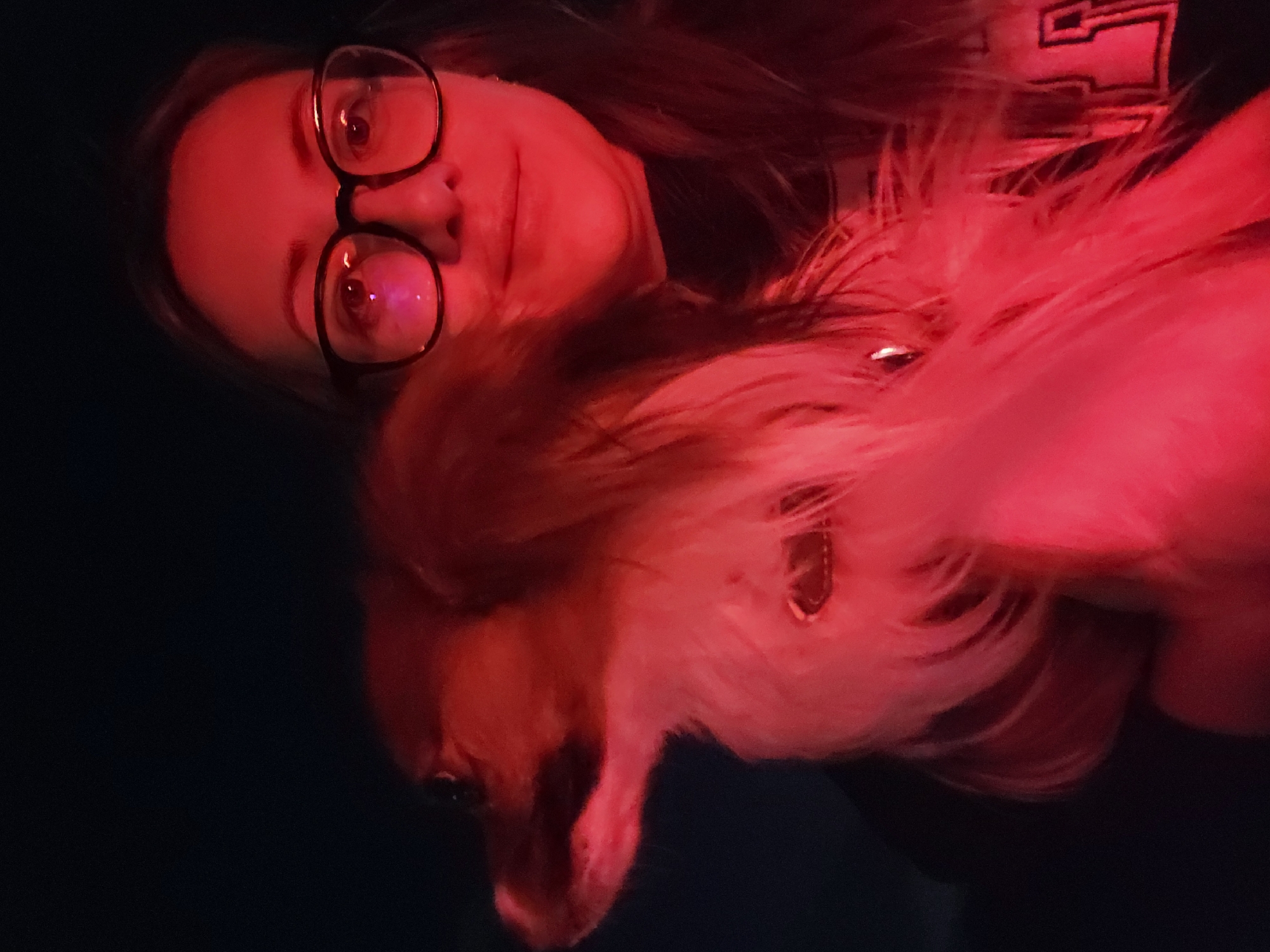 This rotated image captures a close-up of a person and a small dog bathed in intense red light, set against a dark background. The person, appearing to be a woman, is positioned towards the left of the frame, looking directly at the camera with a gentle, slight smile. She wears dark-framed glasses, which reflect the prominent red light, and has visible dark hair. On her dark clothing, near her right shoulder, a white or light-colored emblem resembling a stylized letter 'E' or part of a logo is discernible.
Cuddled beside her, in the center-right of the frame, is a fluffy, long-haired dog, appearing light-colored (possibly white and tan), with its head visible. The dog seems calm and is positioned closely to the person, possibly resting on her chest or lap. A collar or tag is visible around the dog's neck.
The pervasive red lighting dominates the scene, creating a dramatic and intimate mood, suggesting a dimly lit indoor setting or evening time. The dark background emphasizes the subjects illuminated by the artificial light. The image conveys a moment of quiet companionship and affection between the person and their pet. The setting is Mojanovići, Montenegro.
