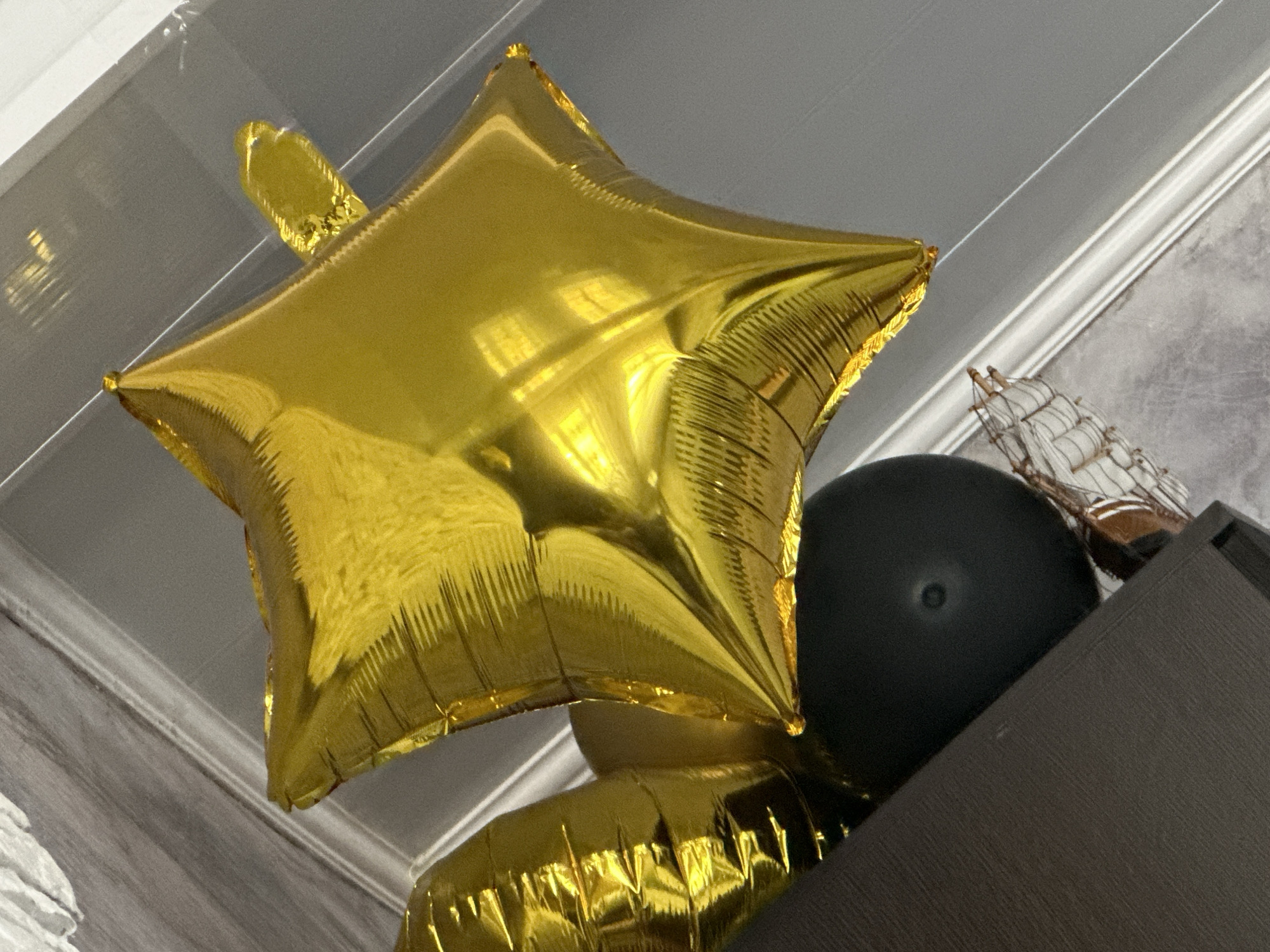 The image shows a close-up, low-angle shot of several decorative items. A large, gold, star-shaped foil balloon dominates the foreground. A black balloon is positioned behind and to the right of the star balloon. In the upper right corner of the frame, there's a small decorative model of a ship. These items appear to be resting on top of a dark-colored, possibly wooden, cabinet or shelf. The background features a textured wall and a white ceiling with molding. The location is Unknown, Unknown.