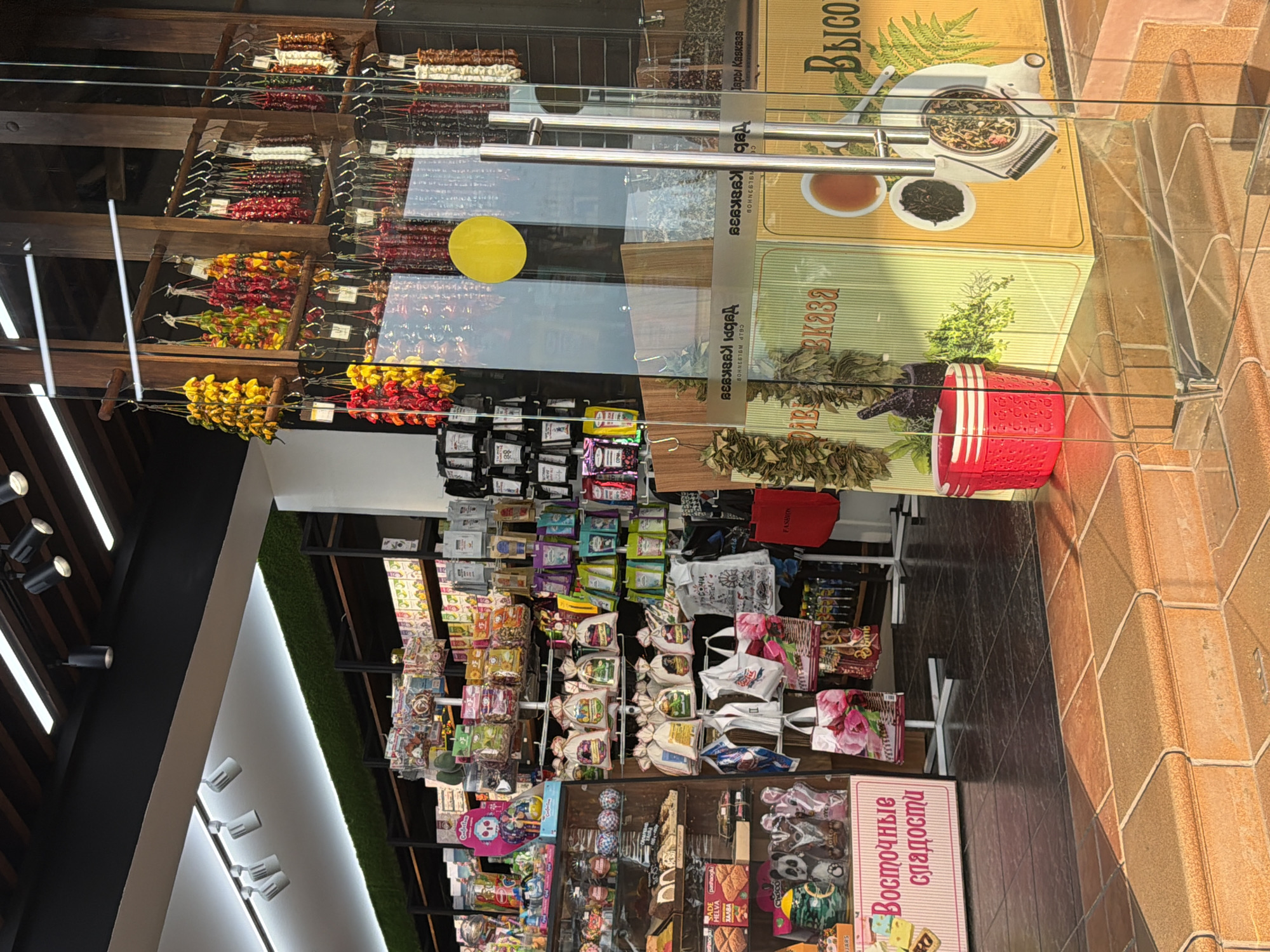 The image shows the entrance to a store that sells dried fruits, nuts, and spices, likely located in Sochi, Russia, given the Cyrillic text visible on signs and packaging. The storefront features large glass doors with metal handles. Inside, shelves are stocked with various goods, including bags of dried goods, packages of sweets, and strings of colorful dried peppers hanging from the ceiling. A red plastic basket is visible on the floor near the entrance, and a pink sign with Cyrillic text "Восточные сладости" (Eastern Sweets) is positioned to the left of the entrance. The lighting suggests it is daytime, with natural light filtering in and interior lights illuminating the merchandise. The overall impression is of a bustling market stall or specialty food shop.