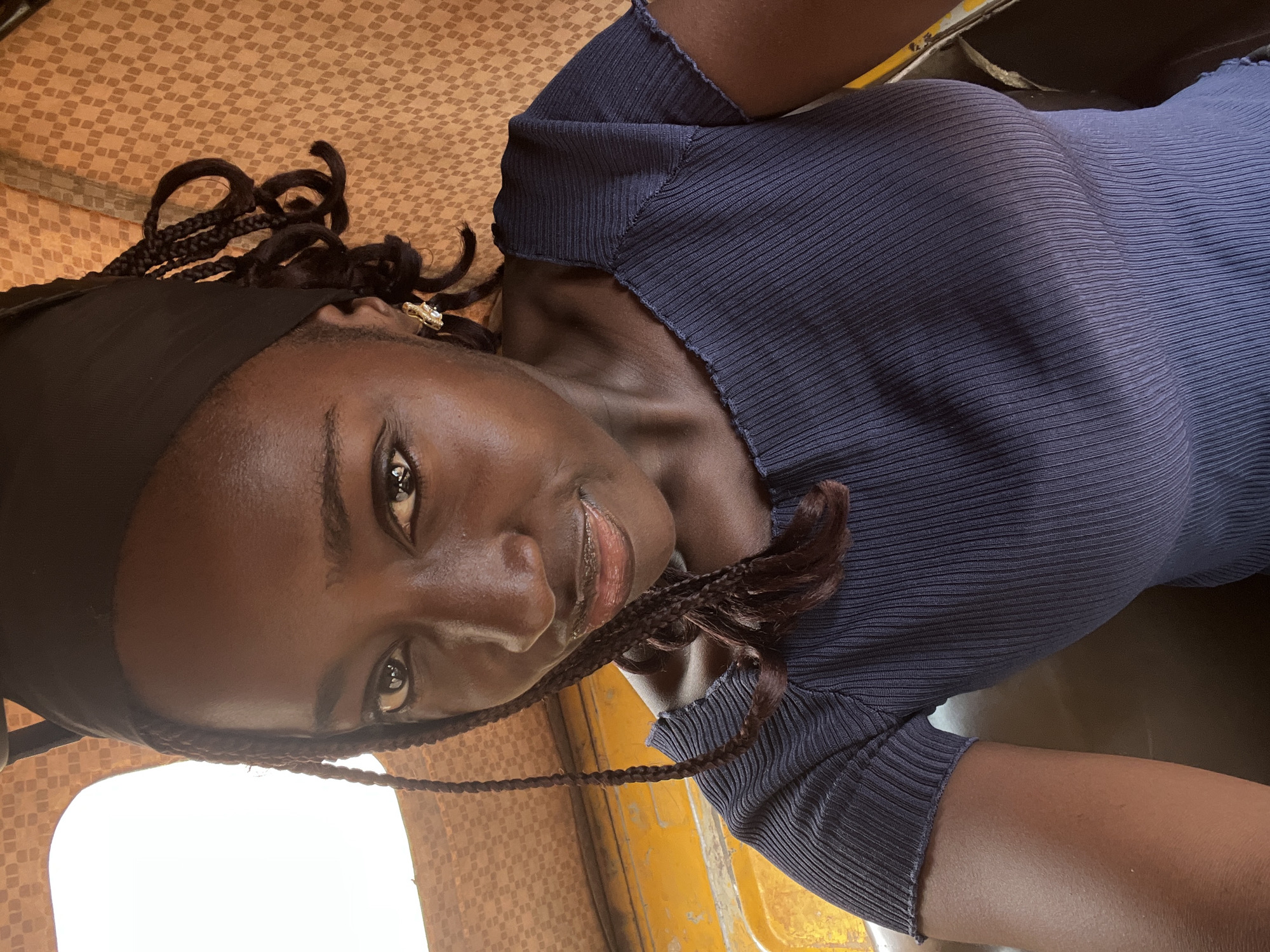A close-up selfie shows a young Black woman with dark skin, braided hair, and a black headscarf, looking towards the camera. She is wearing a dark blue, ribbed, short-sleeved top with a square neckline. She has on hoop earrings. She is in the passenger seat of a vehicle, possibly an auto-rickshaw, with a yellow interior visible. The background is a patterned fabric, likely the vehicle's upholstery. The lighting suggests it is daytime. The scene is indoors, within a vehicle. The location is specified as Abuja, Nigeria.