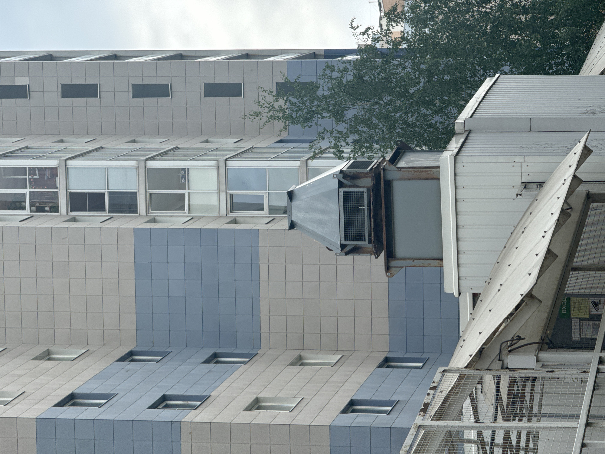 This media file displays a modern urban or industrial scene, captured with a vertical orientation that implies the original image was rotated 90 degrees clockwise.
The primary subject is a tall building featuring a contemporary facade, characterized by a grid of alternating light gray and blue-gray square tiles. The upper section of the building showcases rows of horizontal, rectangular windows, some reflecting the exterior and others offering glimpses into the interior. Below these, smaller, recessed square windows are integrated into the tiled pattern, adding visual depth.
In the mid-ground, a lush green tree partially obscures the building's facade, introducing a natural element to the structured environment. In the foreground, positioned on the right side, is an industrial or utility structure constructed from corrugated metal panels. This structure includes visible metal framework, grates, and what appears to be an air conditioning or ventilation unit. An angular metal ramp or chute, with a weathered texture and visible bolts, extends from this structure towards the bottom right of the frame.
The scene is illuminated by soft, diffused light, suggesting daytime under an overcast sky, as there are no harsh shadows. No human activity or specific events are depicted. On a green sign attached to the industrial structure, partially visible Russian text reads "ВХОД", which translates to "ENTRANCE". The overall impression is that of an architectural and industrial landscape in Saint Petersburg, Russia.