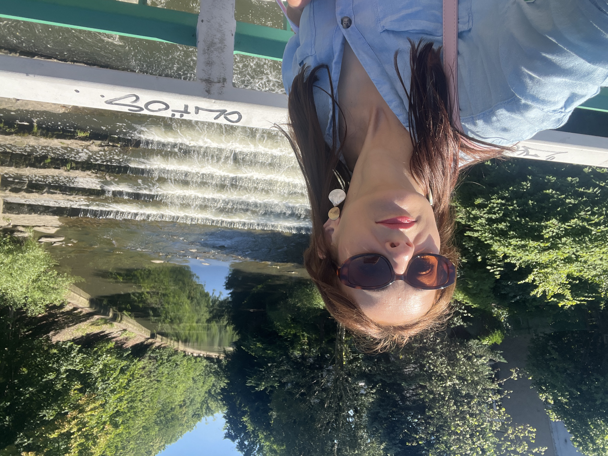 In an outdoor, daylight scene, a woman with long, reddish-brown hair, wearing large, dark sunglasses and a light blue collared shirt, takes a selfie from a bridge. She is positioned in the foreground, with her face angled slightly upwards and to her left, her lips painted with pink lipstick. She wears a pair of large, artistic earrings, one with a round gold element and a white, shell-like pendant. A pink strap, likely from a bag, is visible over her left shoulder. The bridge railing in the foreground is white and appears to have black graffiti. Behind her, a river flows with a series of cascading water steps. The riverbanks are lined with lush green trees and foliage, and the water reflects the clear blue sky and surrounding greenery. In the background, a building is partially visible amongst the trees. The overall impression is a sunny, outdoor setting, possibly a park or riverside area within a city.