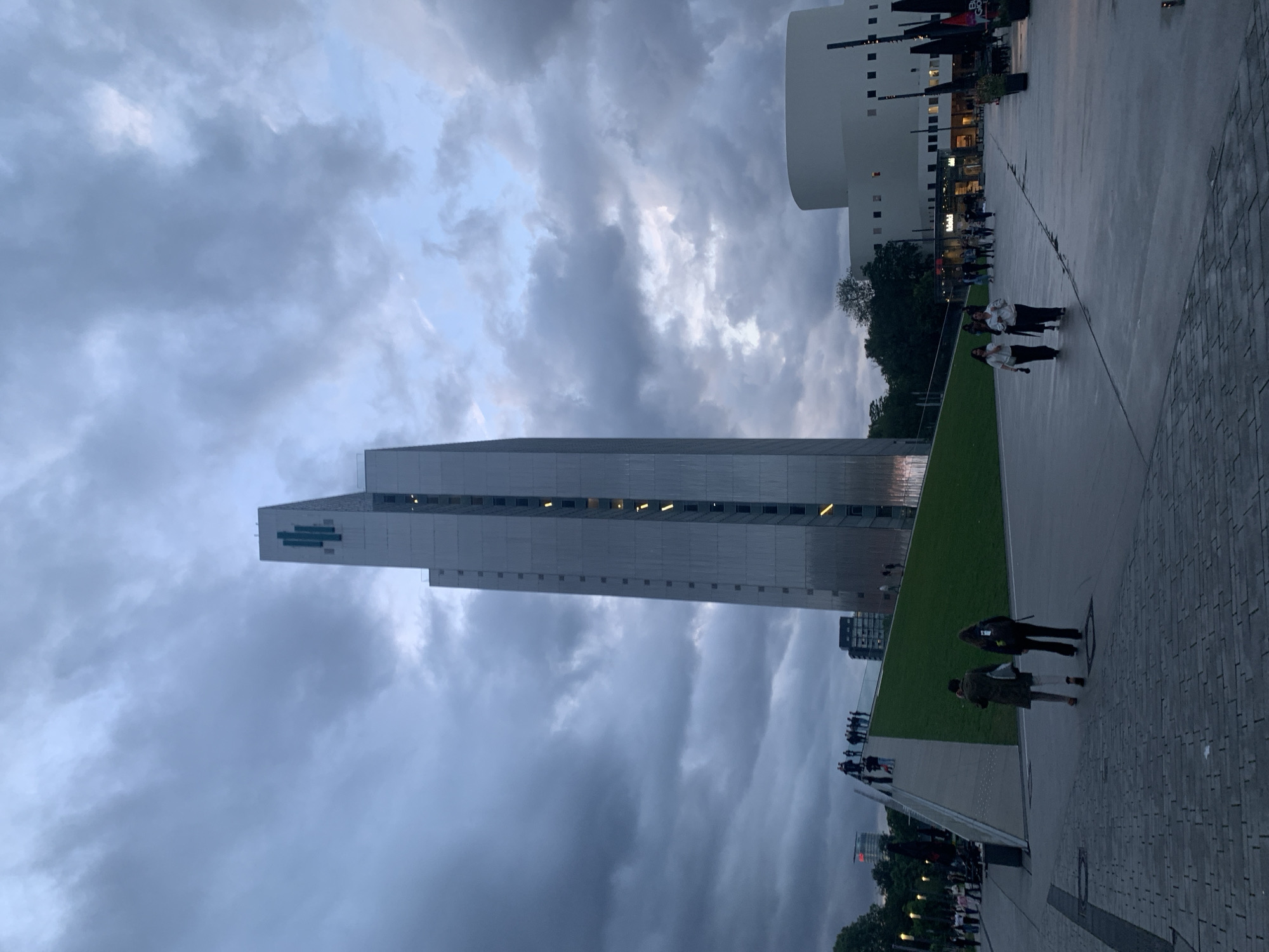 This image captures an urban scene at Wilhelminapier, Rotterdam, Netherlands, under an overcast sky. The focal point is the Maastoren, a tall, slender, modern skyscraper, positioned slightly left of center. Its facade is composed of metallic, possibly corrugated, panels, with a distinctive green, multi-barred Deloitte logo prominently displayed at its top. Vertically aligned windows with internal illumination are visible along the building's height.
To the left of the Maastoren, a large, sloping, triangular structure covered in green grass serves as a pedestrian ramp or elevated plaza. A group of approximately 10-15 individuals is gathered on the upper section of this grassy slope. In the left foreground, two individuals are walking away from the camera on a paved area with a cobblestone-like texture; one wears a dark green coat, and the other wears dark clothing and a backpack.
To the right of the Maastoren, a large, curvilinear, white building, identified as the New Luxor Theater, features a smooth facade with rows of small, rectangular windows. Adjacent to this building, an outdoor cafe or restaurant area is visible, furnished with multiple dark, closed umbrellas or awnings and several planters. In the right mid-ground, two individuals walk towards the camera on a smooth concrete plaza area; both are wearing light-colored tops and dark trousers. Additional people are dispersed throughout the plaza and near the cafe area.
Trees are visible on the far left and behind the New Luxor Theater. The sky is predominantly covered by heavy, dark grey clouds, with patches of lighter blue visible towards the upper right, suggesting either twilight or an overcast daytime. The overall lighting is dim and diffused, with artificial lights visible within the Maastoren and other background structures.
