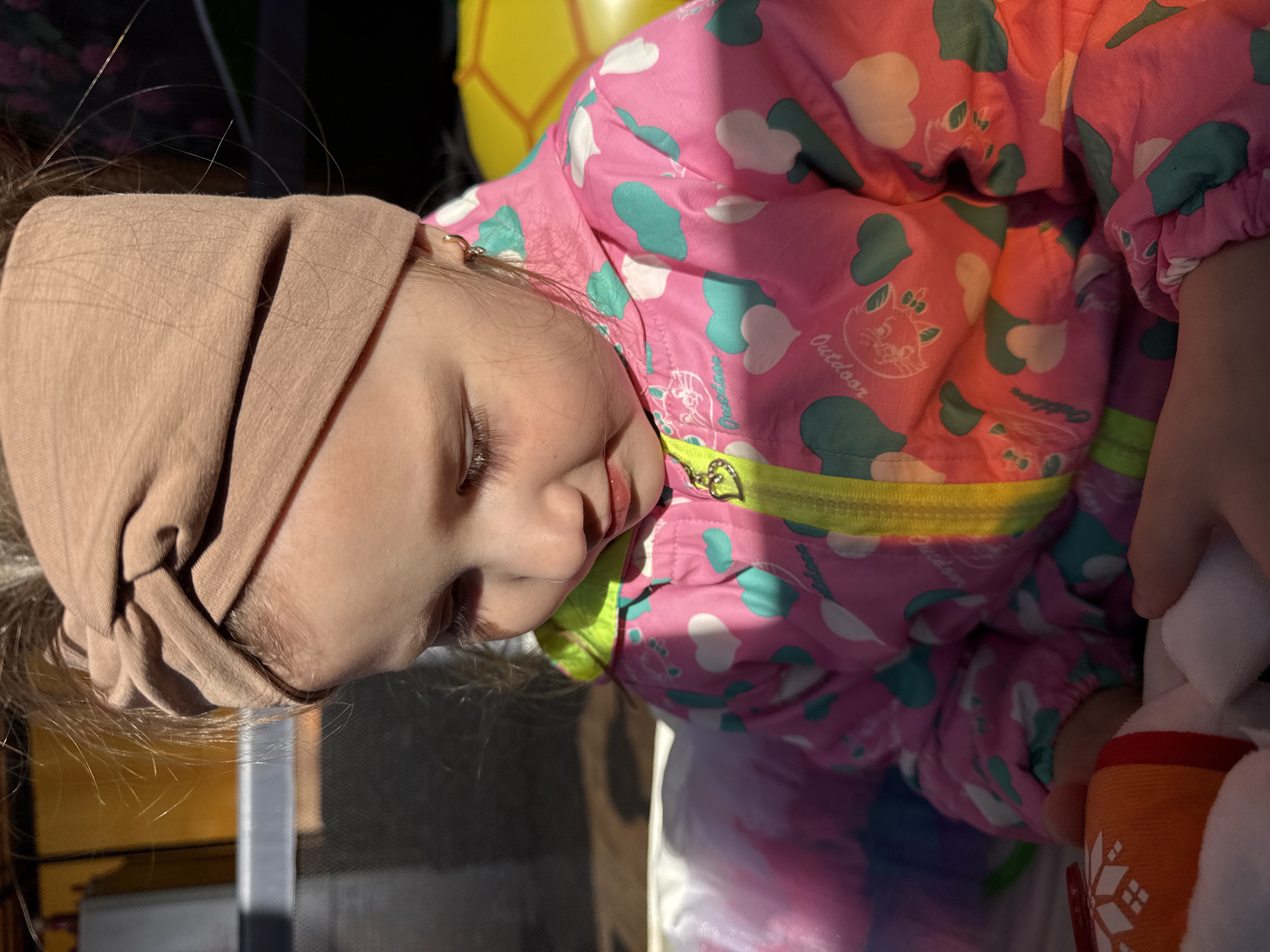 A close-up shot captures a young child with fair skin, appearing to be asleep. The child is wearing a light brown head covering and a pink and green patterned jacket. The jacket features images of white clouds and green hearts, with the word "Outdoor" visible. Part of a yellow balloon is visible in the background. The image seems to have been taken in Кипчак, Russia.