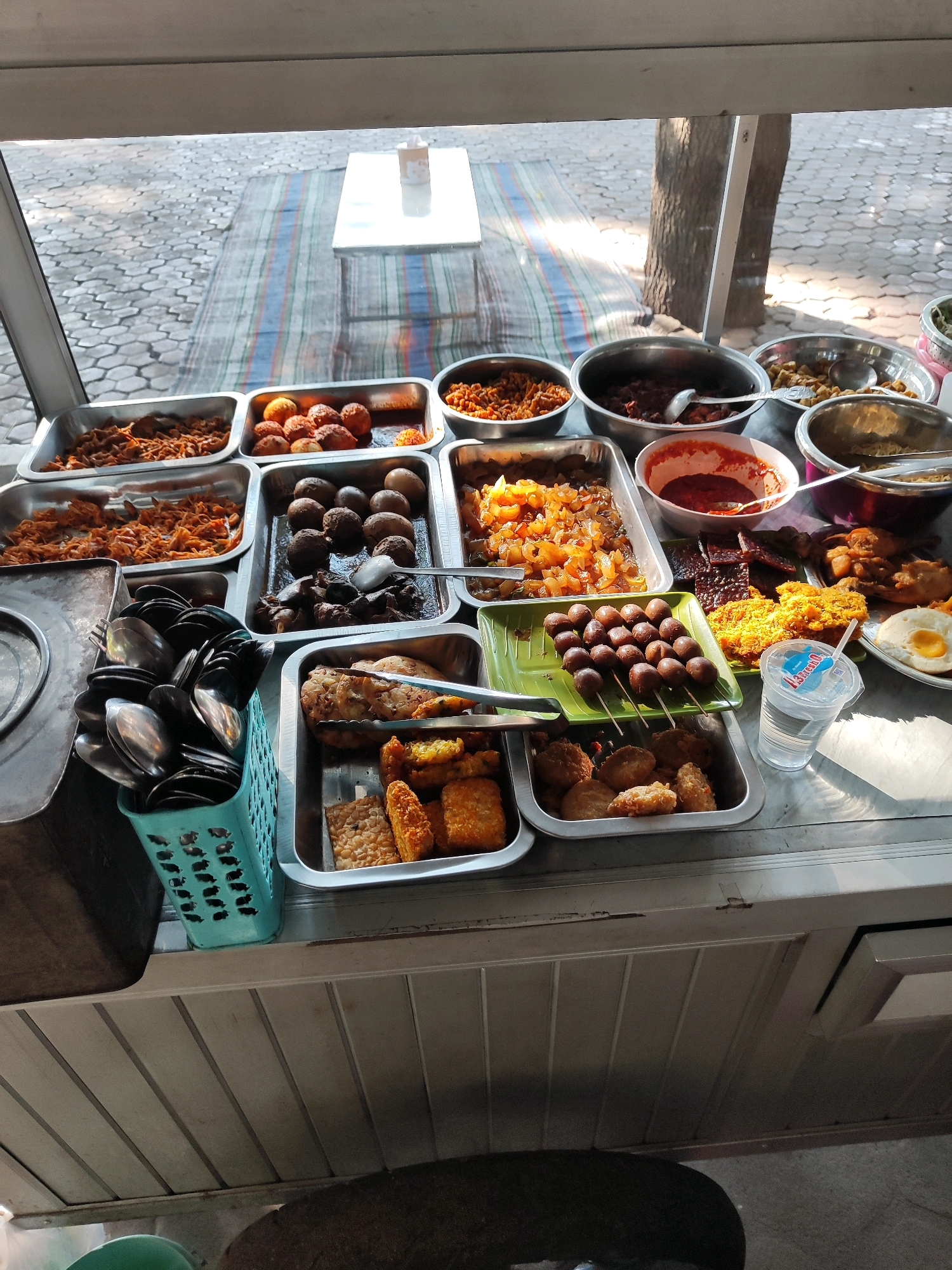 The image depicts a food stall display, likely a street food vendor, in the city of Bondowoso, Indonesia. The stall is set up outdoors, with a tiled ground visible through a window or open front, suggesting a public space. The display features various Indonesian dishes served in metal trays and bowls. These include fried items like tempeh and chicken, meatballs on skewers, seasoned rice, and some kind of stew or curry. There is also a plastic cup with a beverage and a straw, and a basket filled with spoons and other cutlery. The overall impression is one of a bustling local eatery offering a diverse range of ready-to-eat meals.