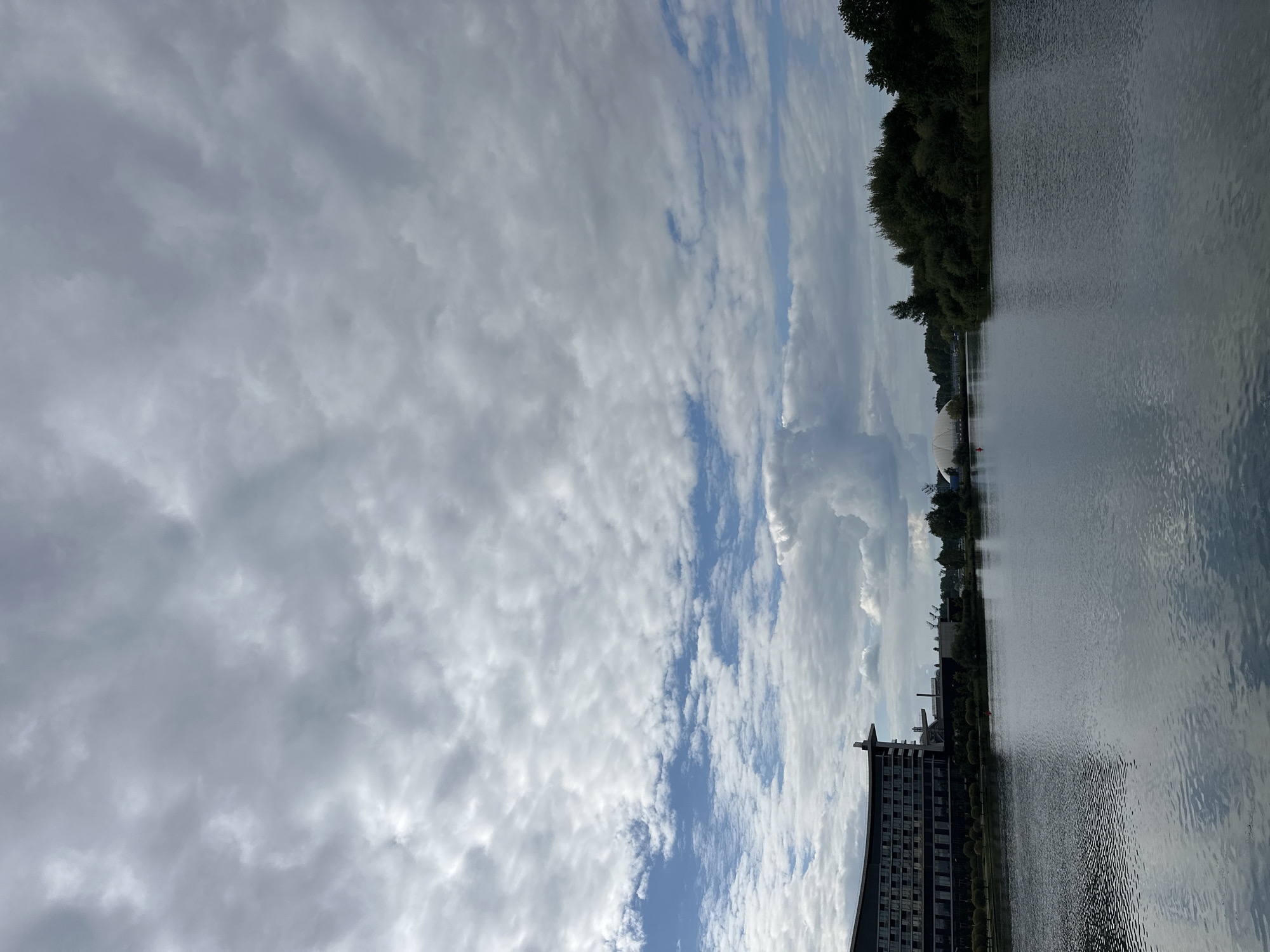 This is a wide-angle, eye-level shot of a calm body of water, likely a reservoir or a large pond, in Minsk, Belarus. The water's surface is smooth with subtle ripples reflecting the overcast sky. In the foreground, the water fills the lower half of the frame. On the left side of the image, a modern, multi-story building with dark facades and numerous windows is visible. Behind it, industrial structures and trees are present. The right side of the frame is dominated by a dense line of green trees. In the distance, across the water, a large, white, domed structure is partially visible, along with other buildings and more trees. The sky is heavily clouded with varying shades of gray and white, with patches of blue sky peeking through. No people or animals are visible in the scene. The overall impression is one of a somewhat urbanized, yet natural, landscape under a cloudy sky.