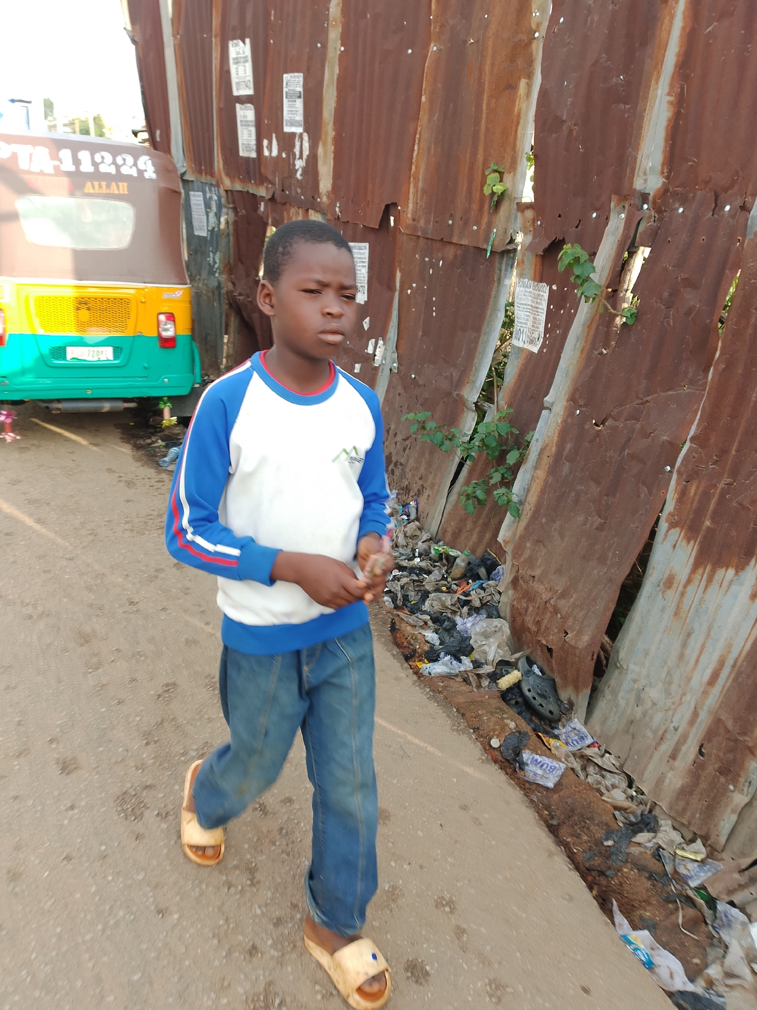 In Jos, Nigeria, a young boy walks on a dusty, unpaved road. He is wearing a white long-sleeved shirt with blue sleeves and trim, and blue jeans. On his feet are light-colored slide sandals. To his left, a brown and yellow auto-rickshaw, identified with "PTA-11224" and "ALLAH" written on its side, is parked. A rusty corrugated metal fence, adorned with faded posters, lines the right side of the road, separating it from an area filled with scattered refuse, including plastic bags and debris. The environment appears unkempt, with plants growing through gaps in the fence and accumulating waste along the roadside. The boy is looking to his right, with his hands slightly extended and holding a small object.