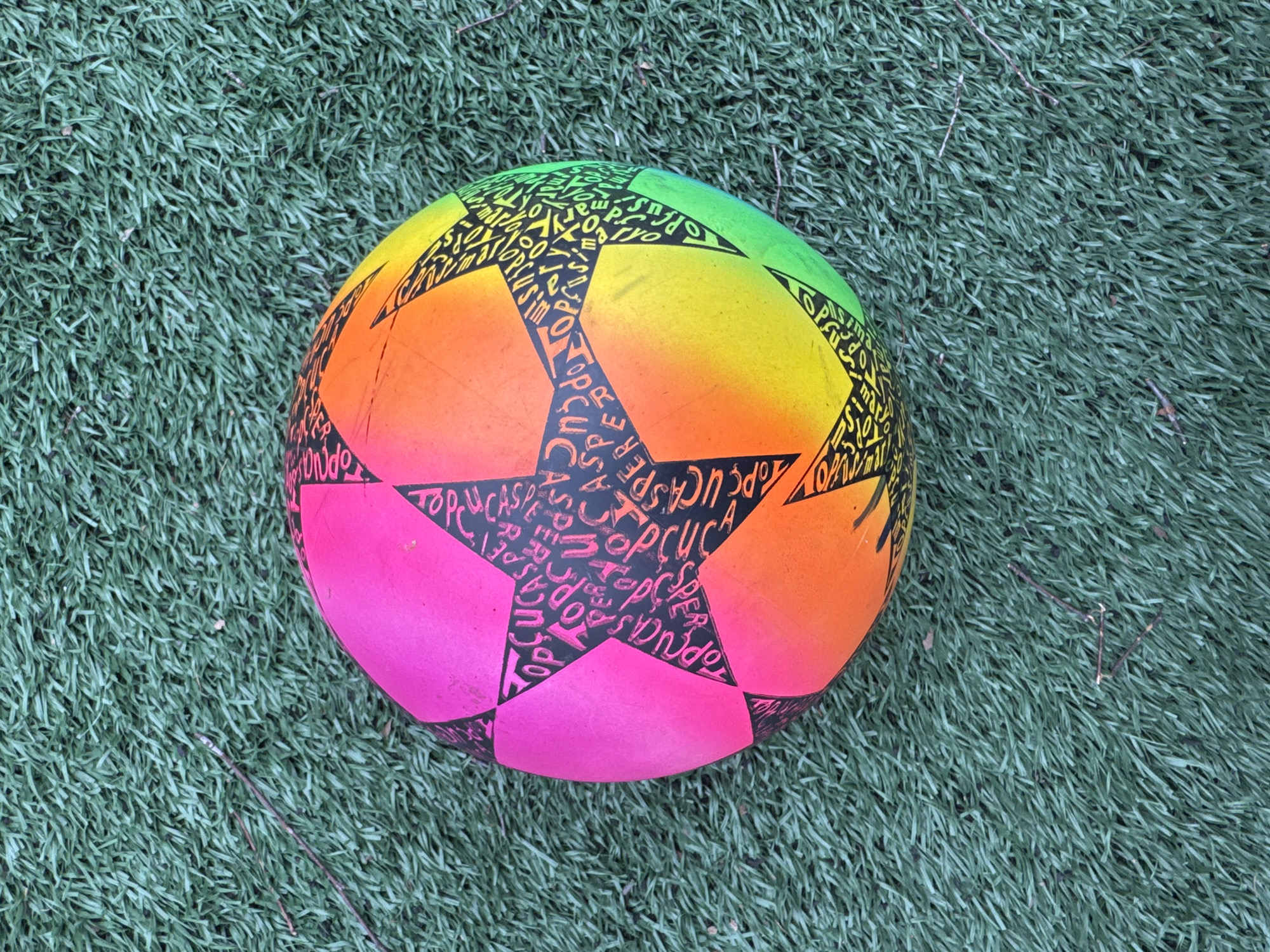A rainbow-colored soccer ball with black star shapes filled with text rests on artificial green grass. The color gradient of the ball transitions from green to yellow to orange to pink. The text within the star shapes appears to be a repeating word or phrase. The artificial grass blades are densely packed and uniformly aligned. There are a few small brown twigs scattered on the grass. The image lacks any indications of being located in Saint Petersburg, Russia.