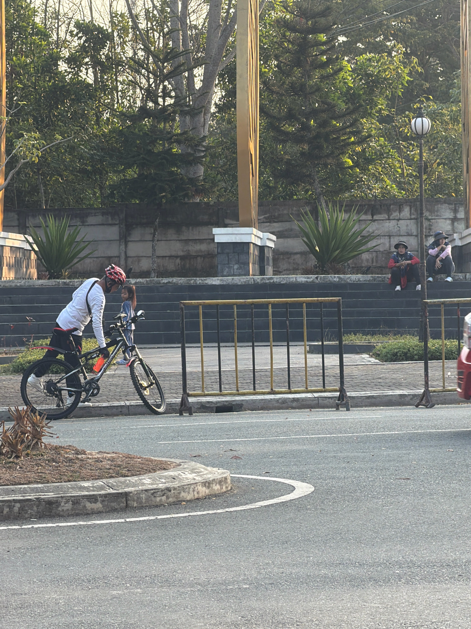 The image captures an outdoor scene in Balikpapan, Indonesia, likely a park or public area with a roadway in the foreground. A man wearing a helmet and a white long-sleeved shirt is leaning over a bicycle, appearing to interact with a young girl standing beside him. The girl is wearing a blue outfit. Behind them, a set of stone stairs leads up to a higher level. Two people, one wearing a red and black jacket and the other in a pink top, are seated on the stairs. The environment features trees, a decorative lamppost, and a concrete wall. A yellow and black metal barrier separates the sidewalk from the road. The road itself has a white lane marker and a curved white line indicating a turn. The lighting suggests it is daytime, possibly late afternoon, with sunlight casting shadows.