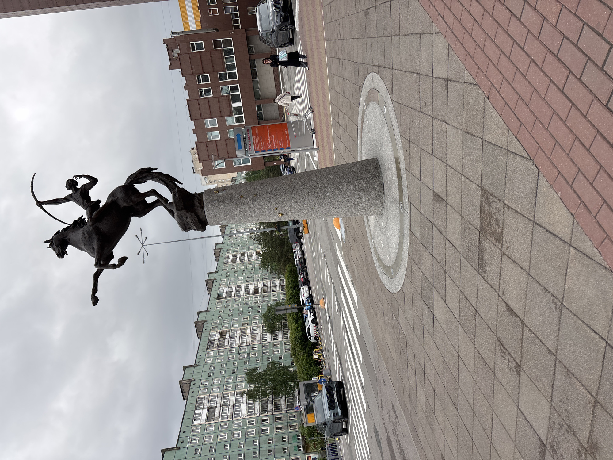 The image depicts the "Horse Tamers" sculpture in Saint Petersburg, Russia. The bronze sculpture features a man on horseback, poised to shoot an arrow. It is mounted on a gray cylindrical pedestal. The sculpture is located on a pedestrian walkway with gray rectangular pavers, bordered by red bricks on one side. A crosswalk is visible in the background, along with parked cars and multi-story buildings with numerous windows. People are walking on the sidewalk near a brown building. The sky is overcast.